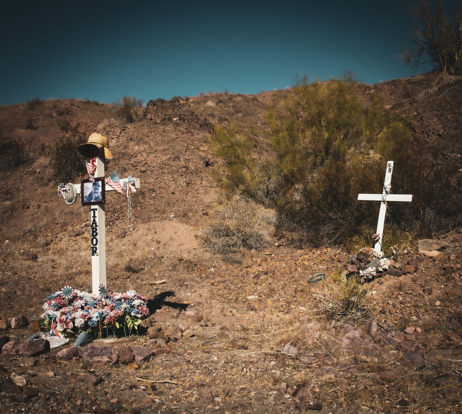 Two roadside memorial crosses with flowers and decorations on a dry desert hillside.