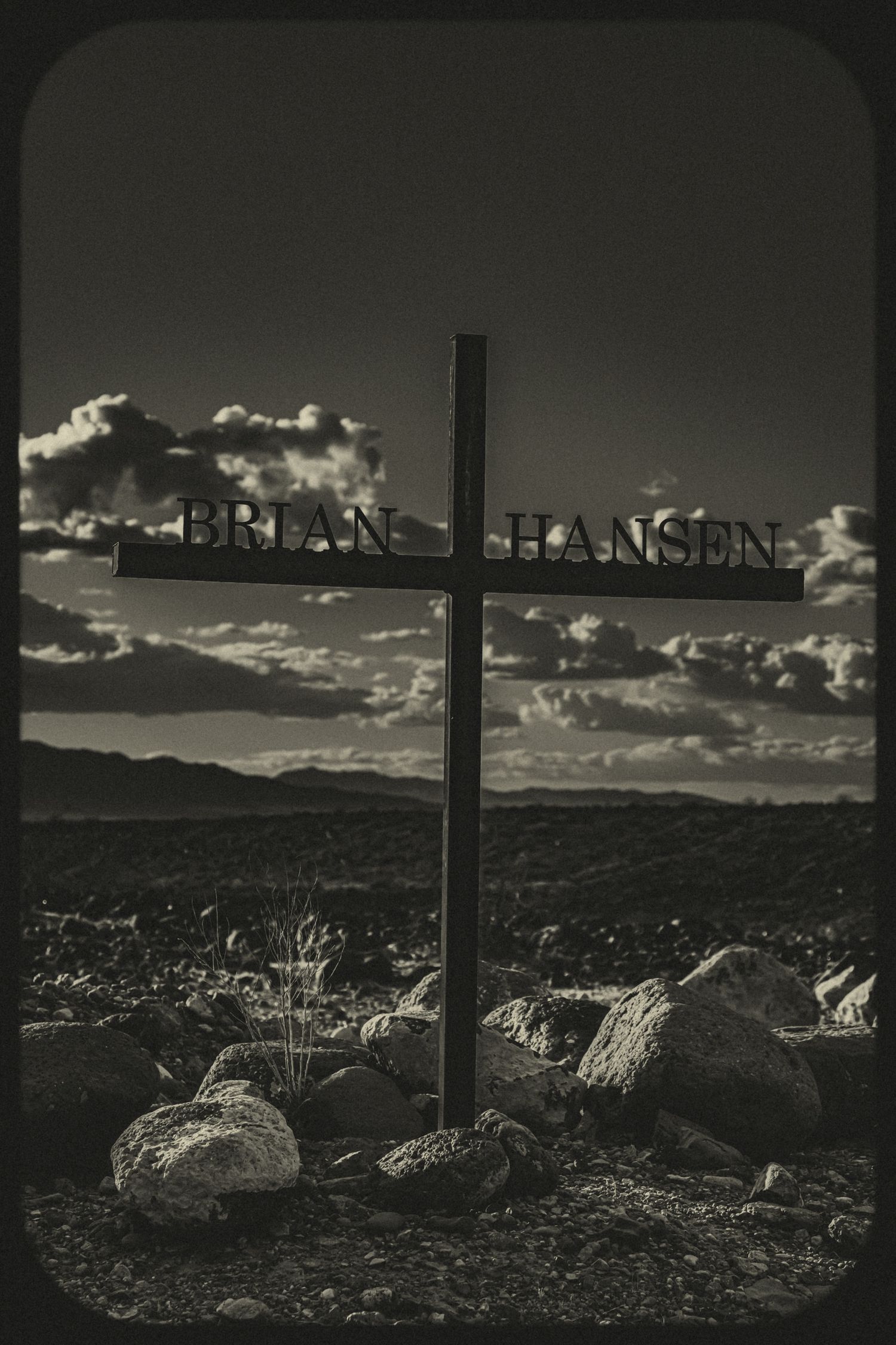 Black and white photo of a roadside memorial cross bearing the name Brian Hansen against a cloudy sky.