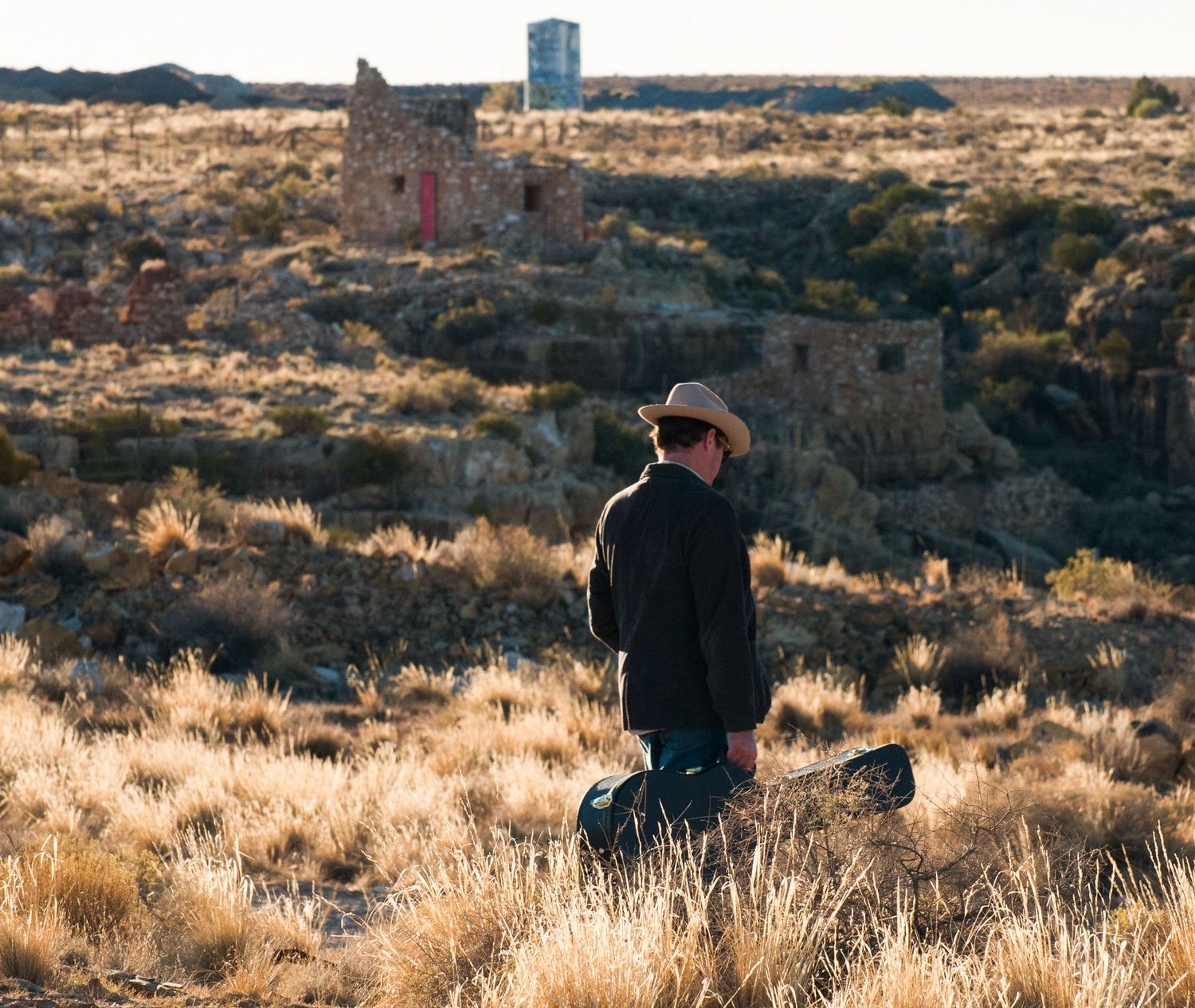 Cowboy in hat and jacket standing in arid scrubland with rustic stone ruins in the background.