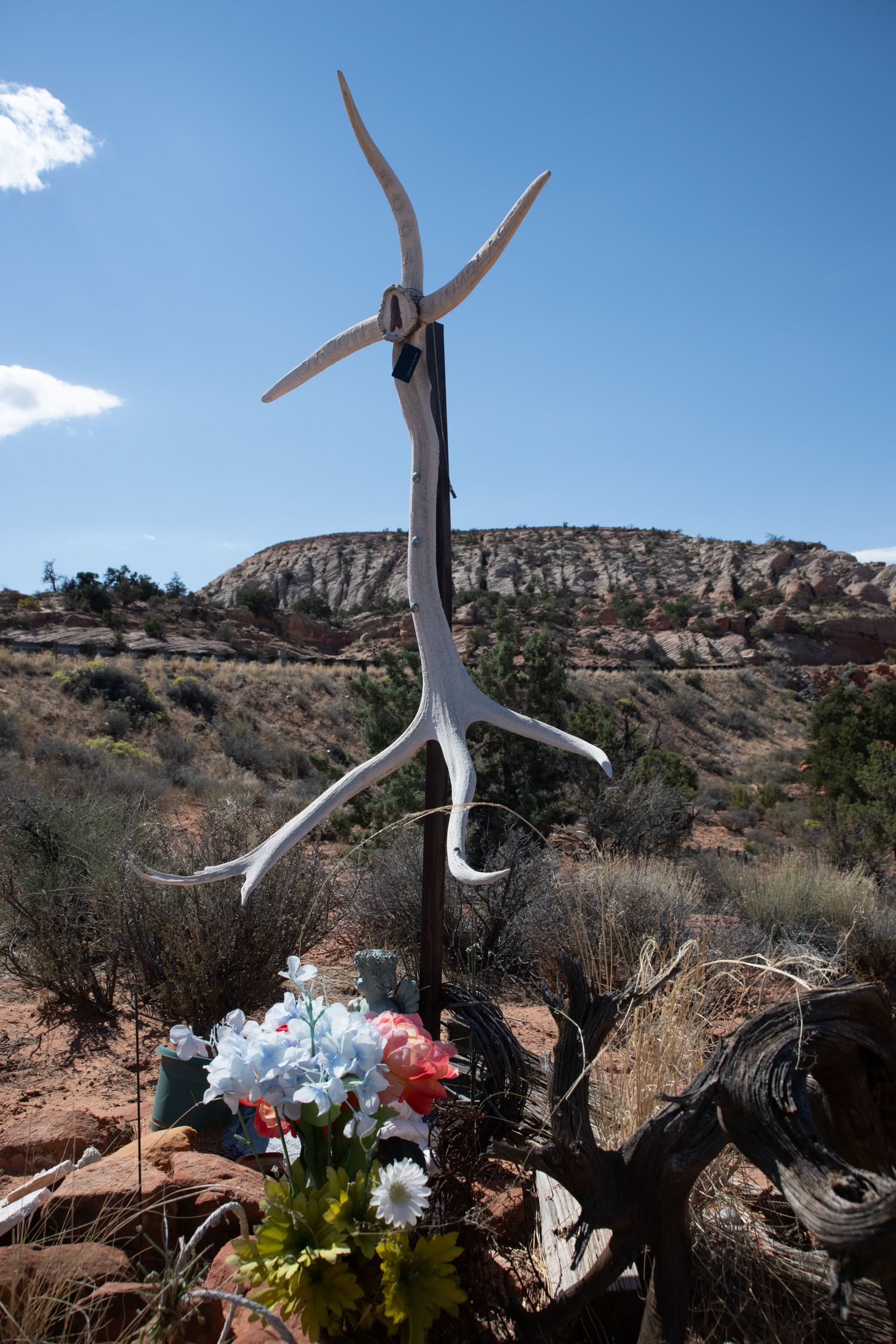 Desert roadside memorial featuring antler cross with colorful flowers against red sandstone cliffs.