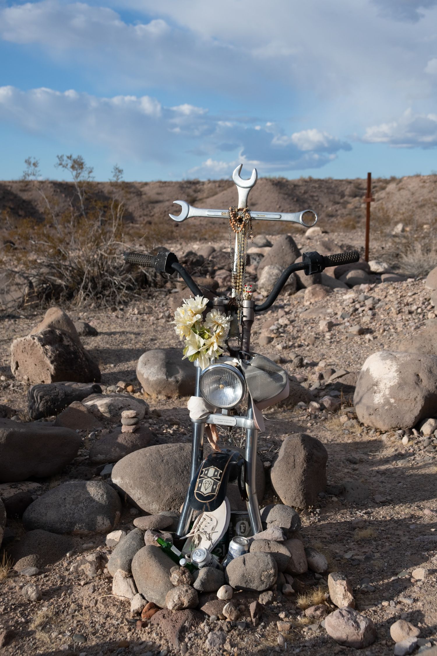 Roadside memorial in desert with motorcycle headlight and wrench cross surrounded by rocks.