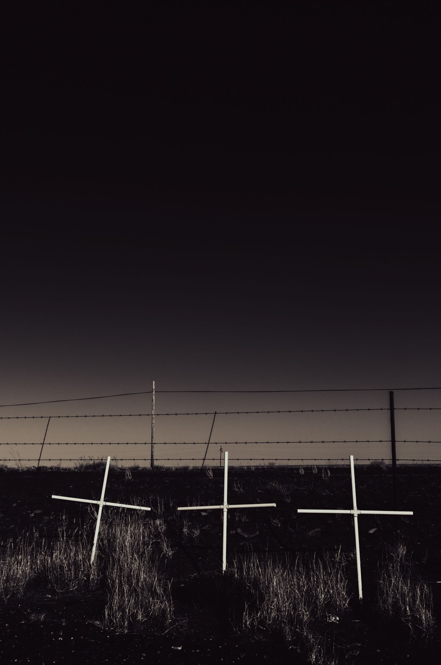 Three white crosses standing in a dark field behind barbed wire fence at night.
