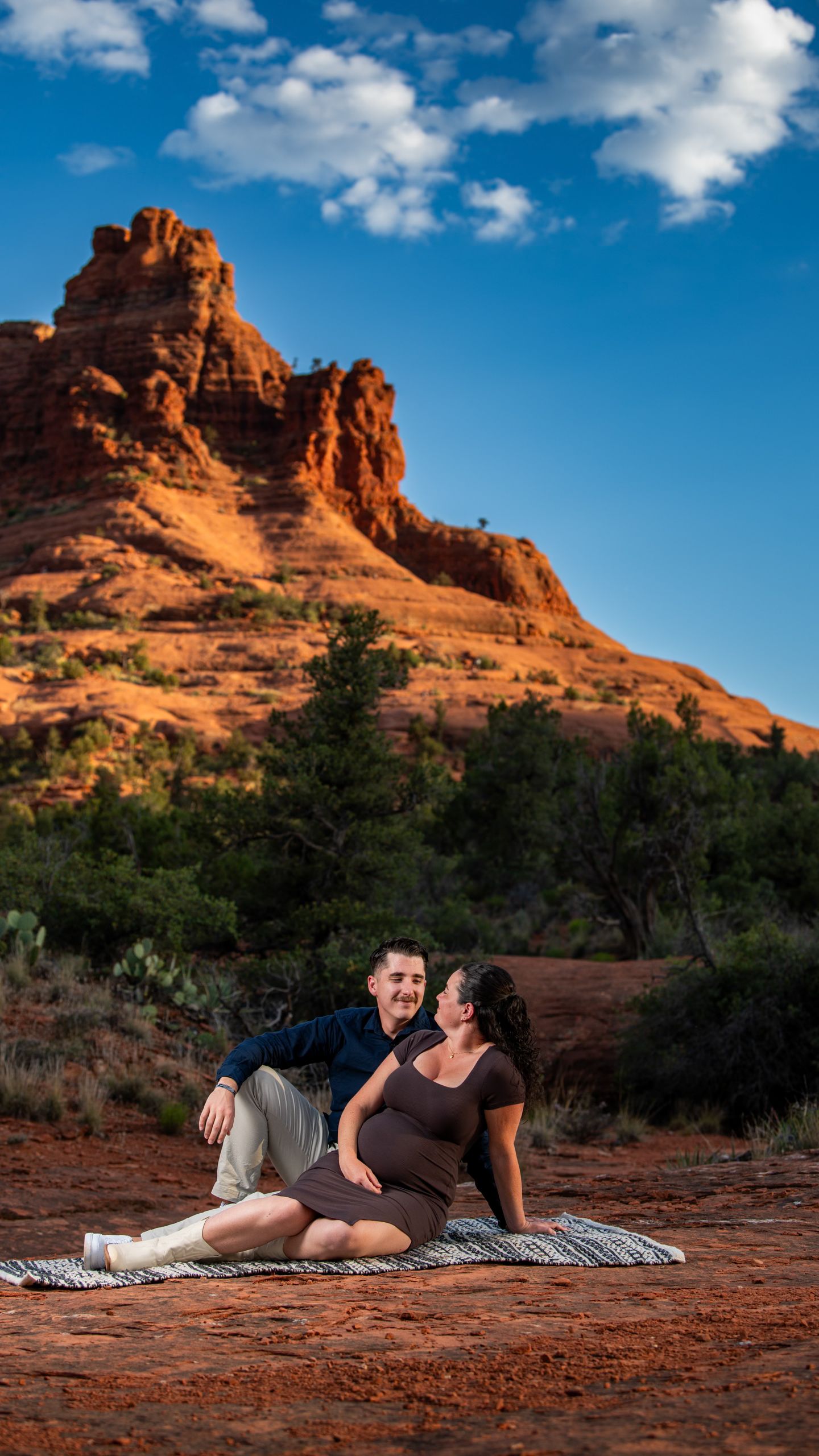 Expecting couple sitting on blanket in Sedona with glowing red rock butte under blue cloudy sky.
