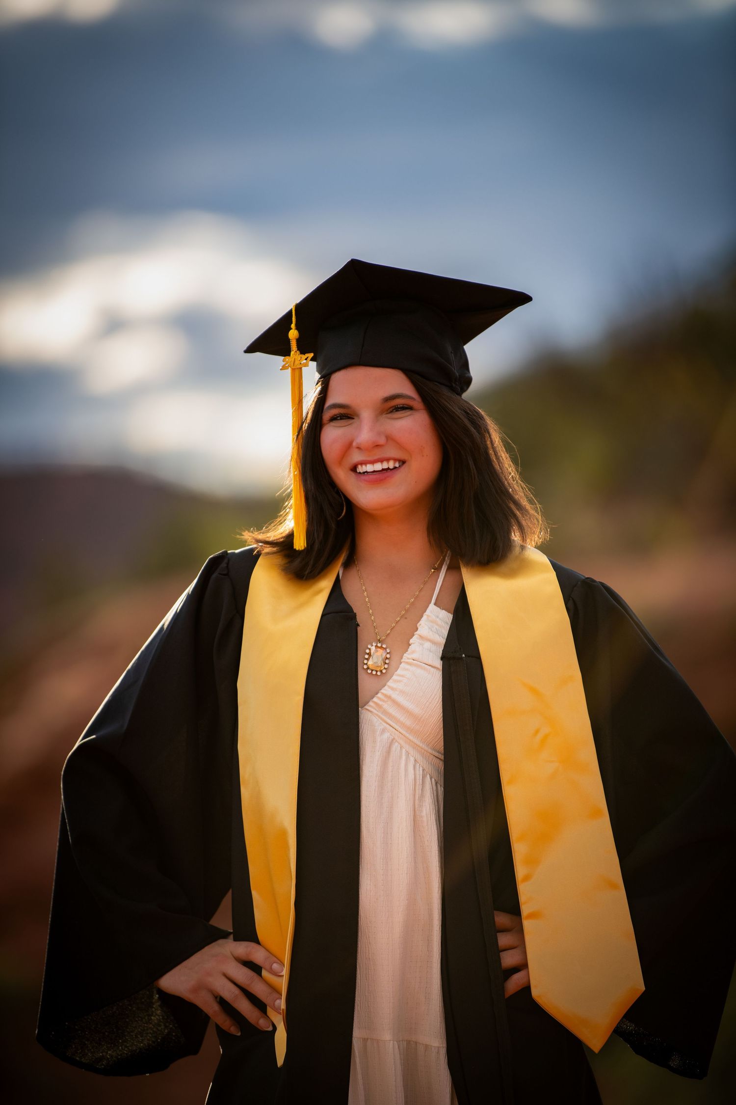 Smiling young woman in black graduation cap and gown with gold stole poses outdoors at golden hour.