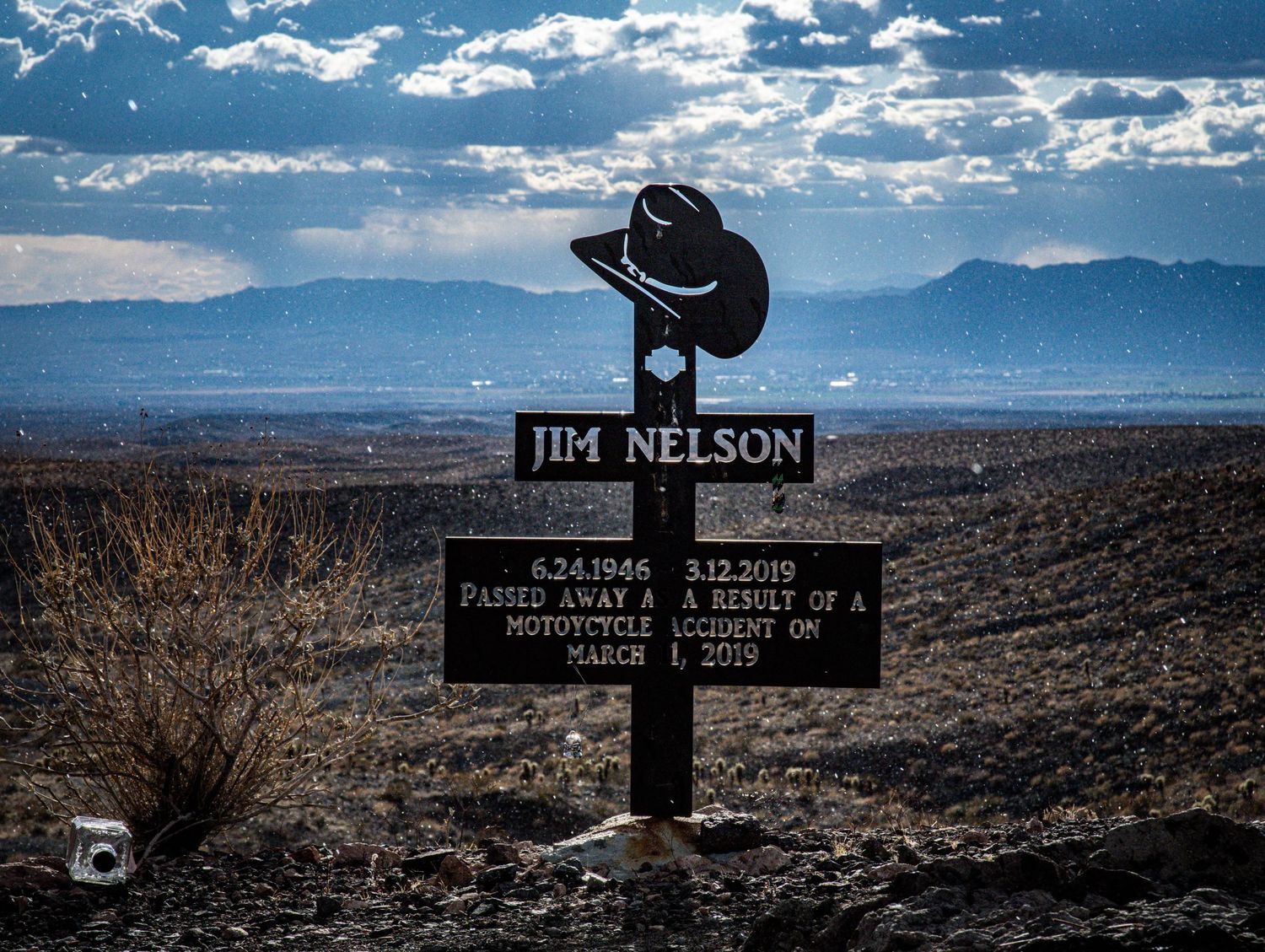 Roadside memorial cross for Jim Nelson, a motorcycle accident victim, with cowboy hat silhouette against desert landscape.