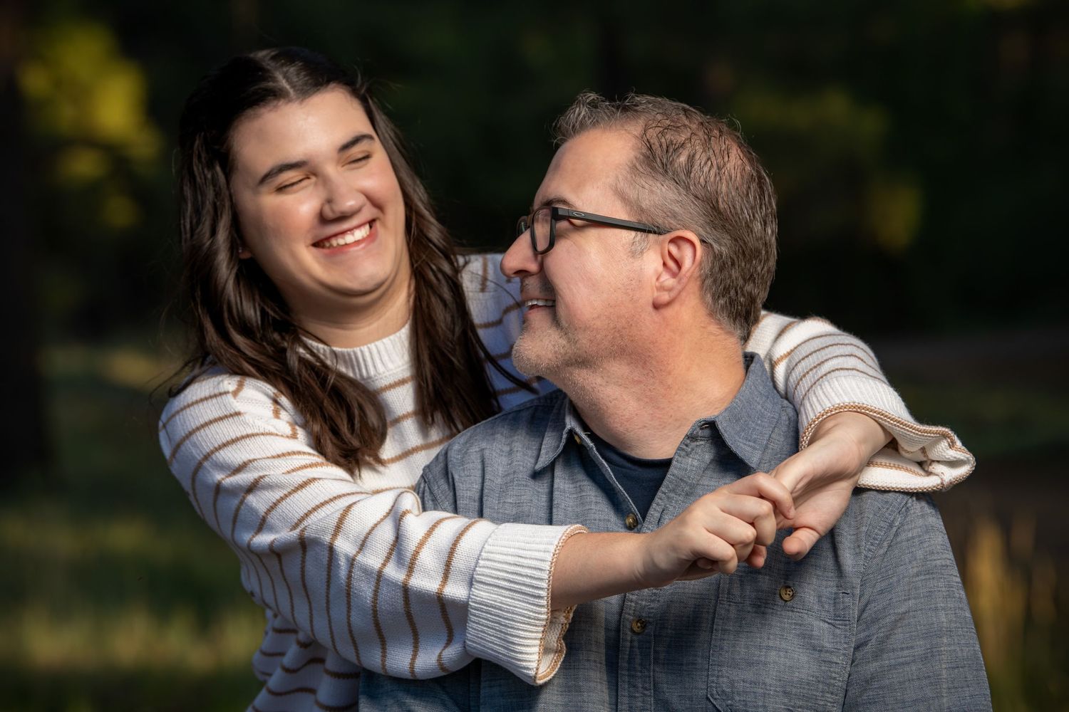 Young woman hugs a smiling older man from behind in a warmly lit outdoor autumn park setting.