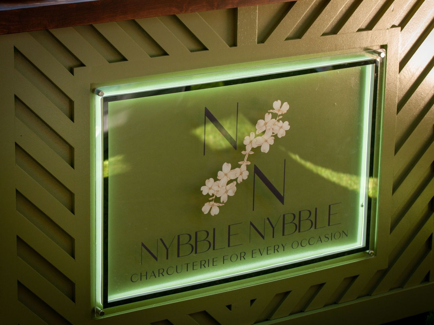Illuminated Nybble Nybble charcuterie sign with floral design on green lattice wall.