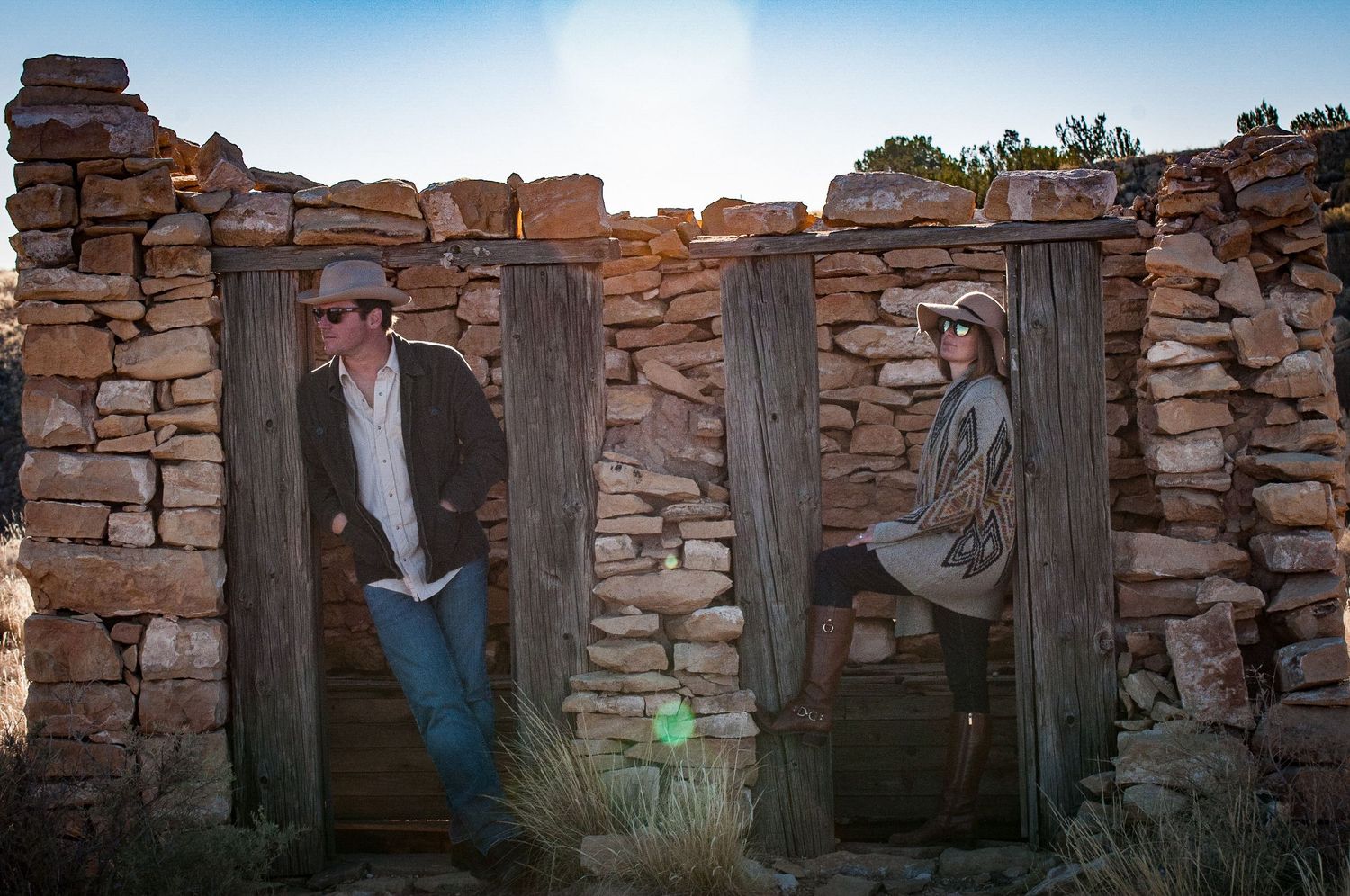 Man and woman posing in separate doorways of a rustic stone wall structure at sunset in the desert.