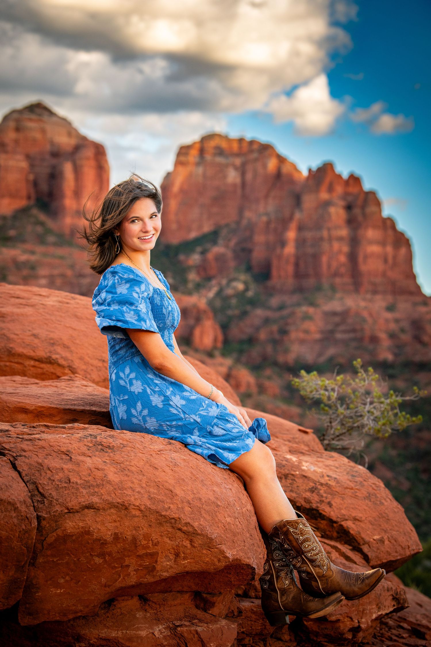 Woman in blue floral dress and cowboy boots sitting on red rocks in Sedona, Arizona with dramatic buttes behind her.