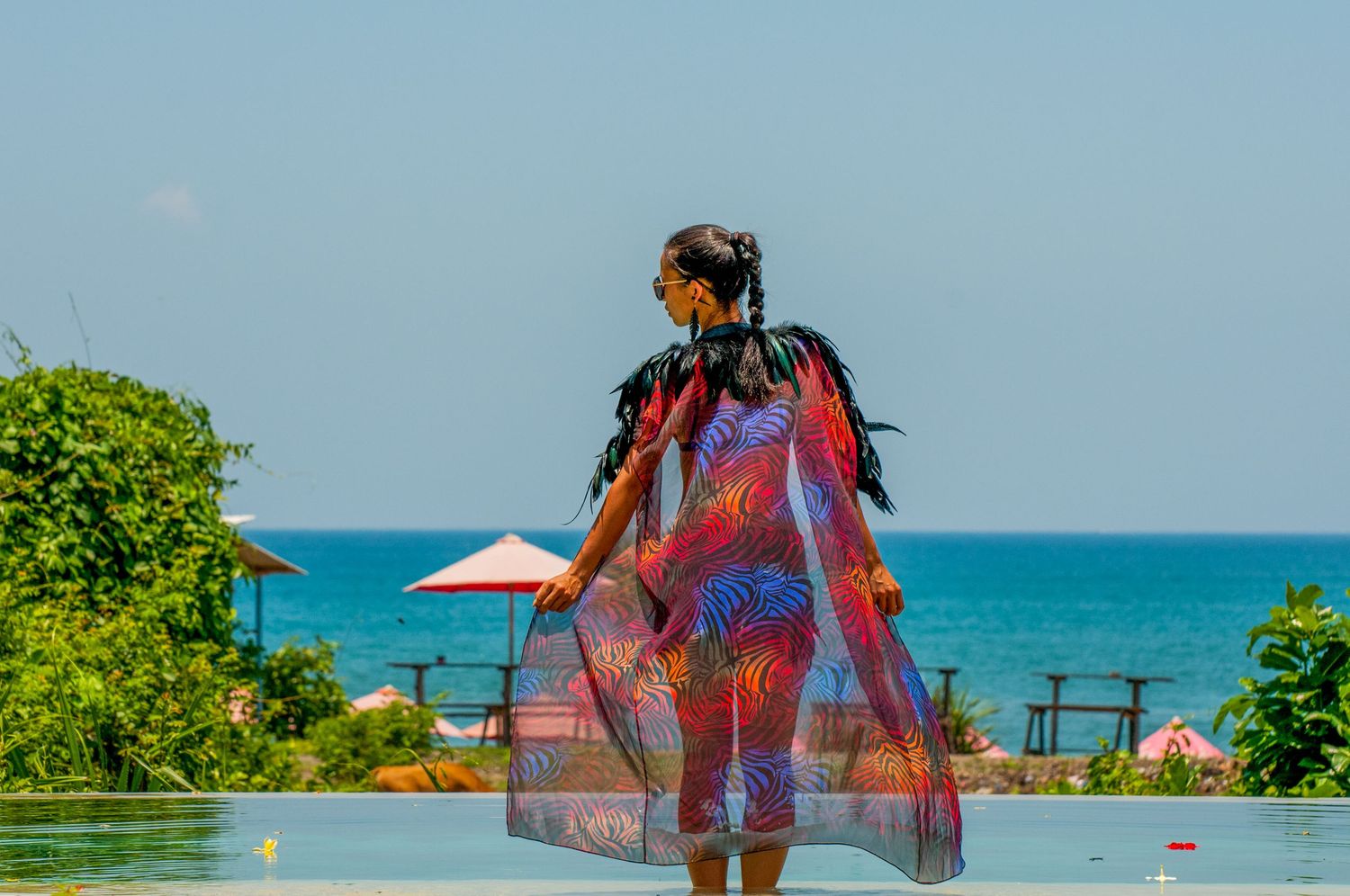 Woman in colorful sheer cover-up standing by an infinity pool overlooking a tropical ocean view.