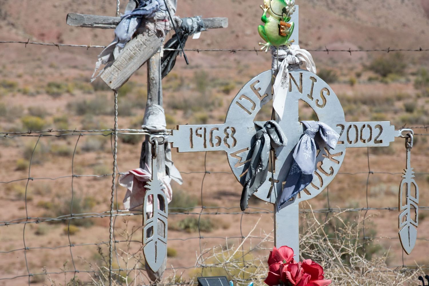 Close-up of roadside memorial cross for Dennis 1968-2002 with feathers and flowers in desert.
