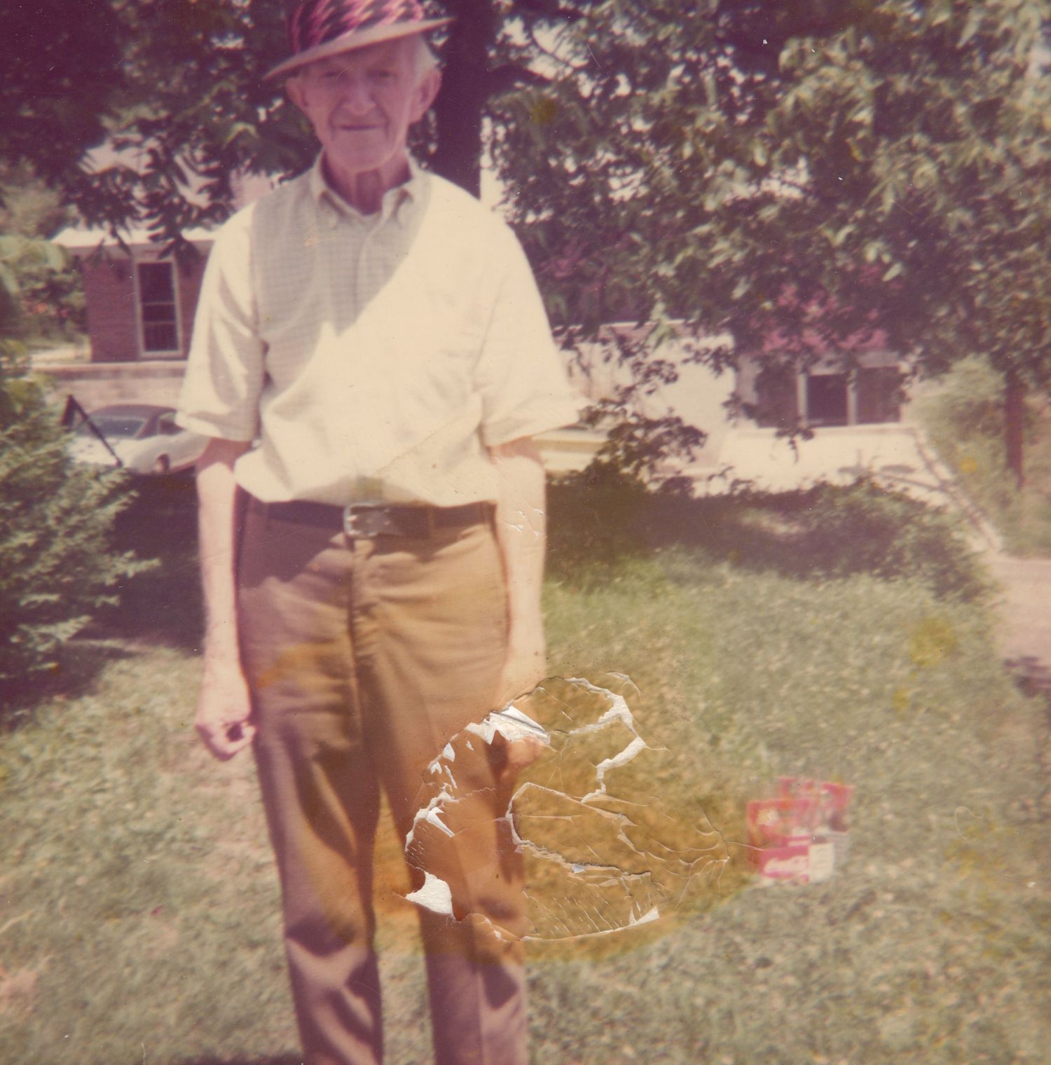 Elderly man standing in a green yard holding a decorative item, wearing a hat and casual clothes near a house.