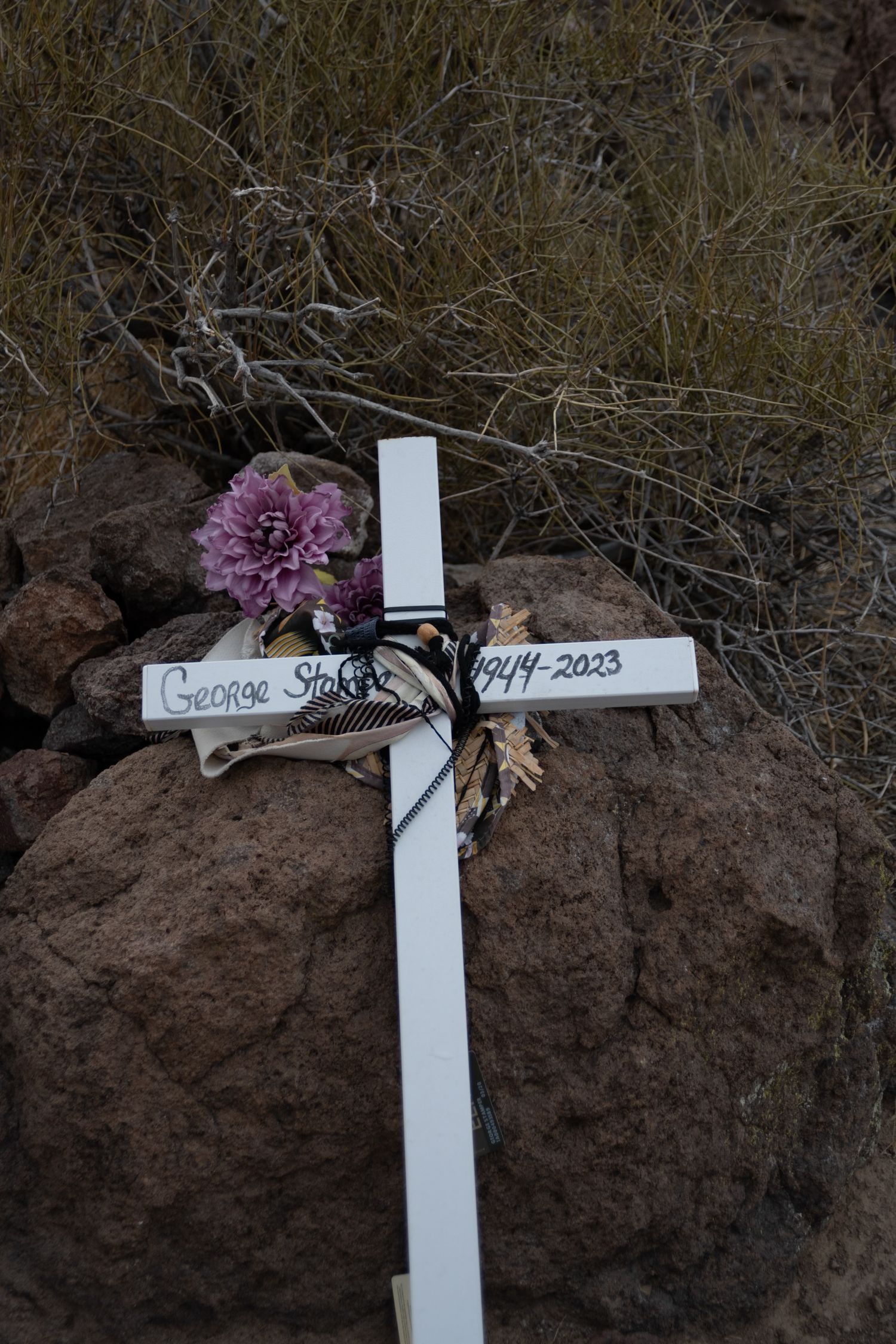 White memorial cross for George Stakes 1944-2023 adorned with purple flowers on rocky desert terrain.