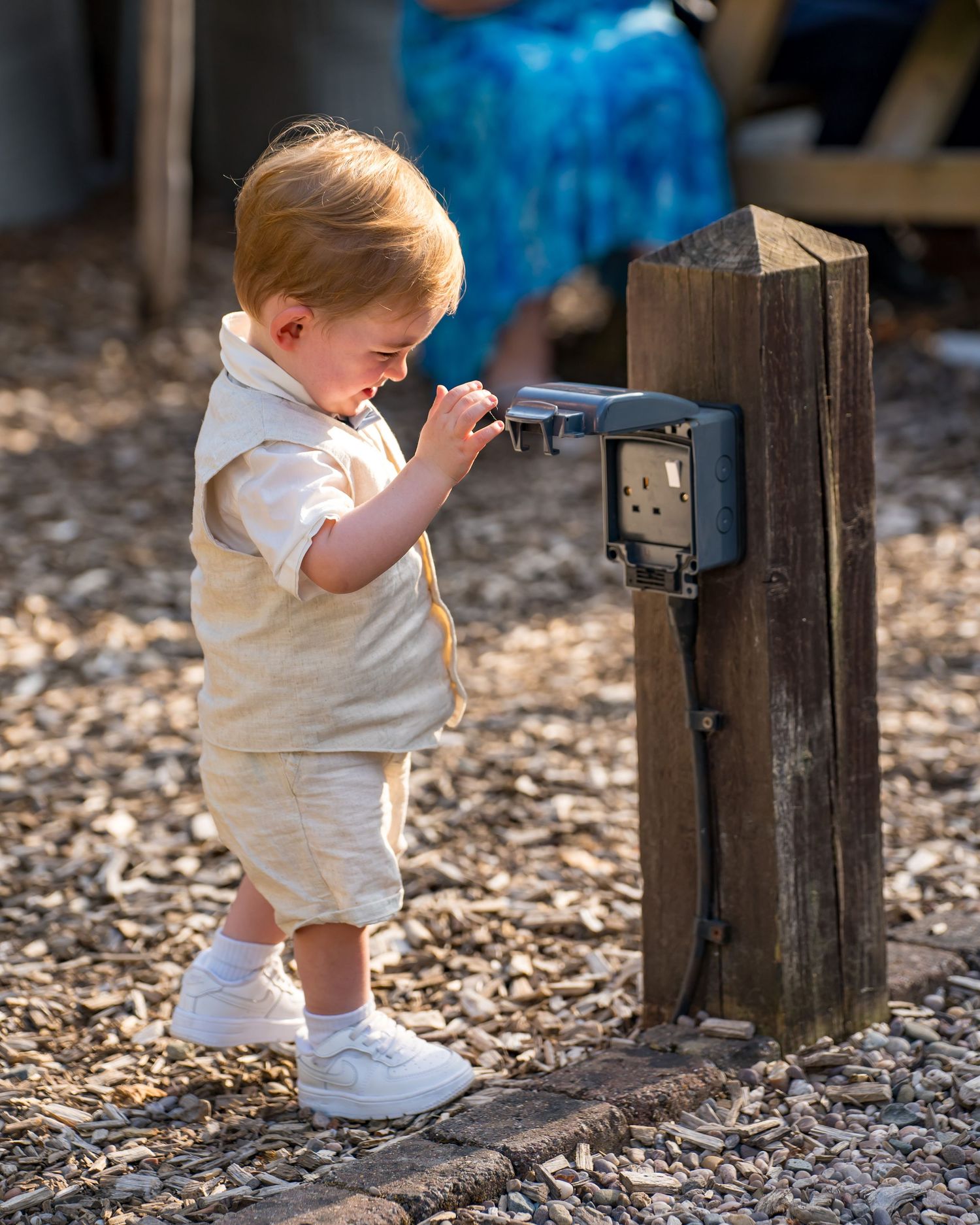 A toddler in a cream outfit explores a wooden post with an electrical box on a gravel path outdoors.