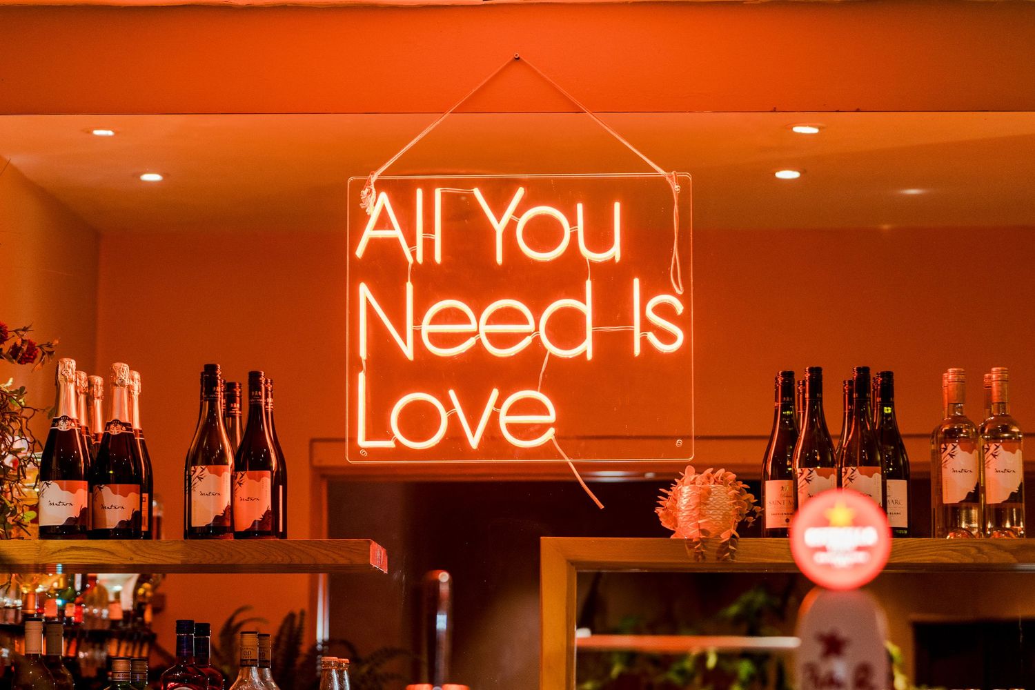 Neon sign reading All You Need Is Love glows orange above bar bottles at wedding venue.
