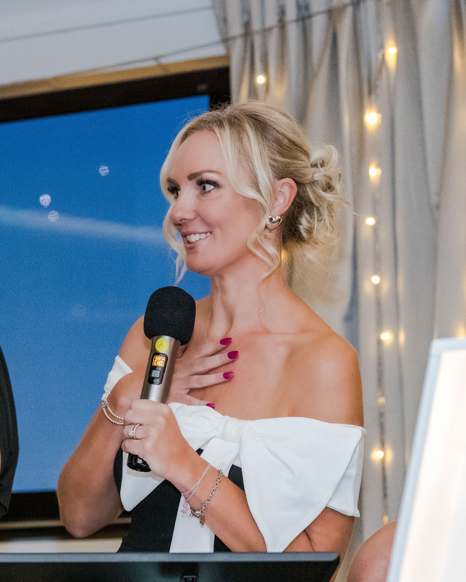 Elegant blonde woman in off-shoulder white dress speaking into microphone at a formal event.