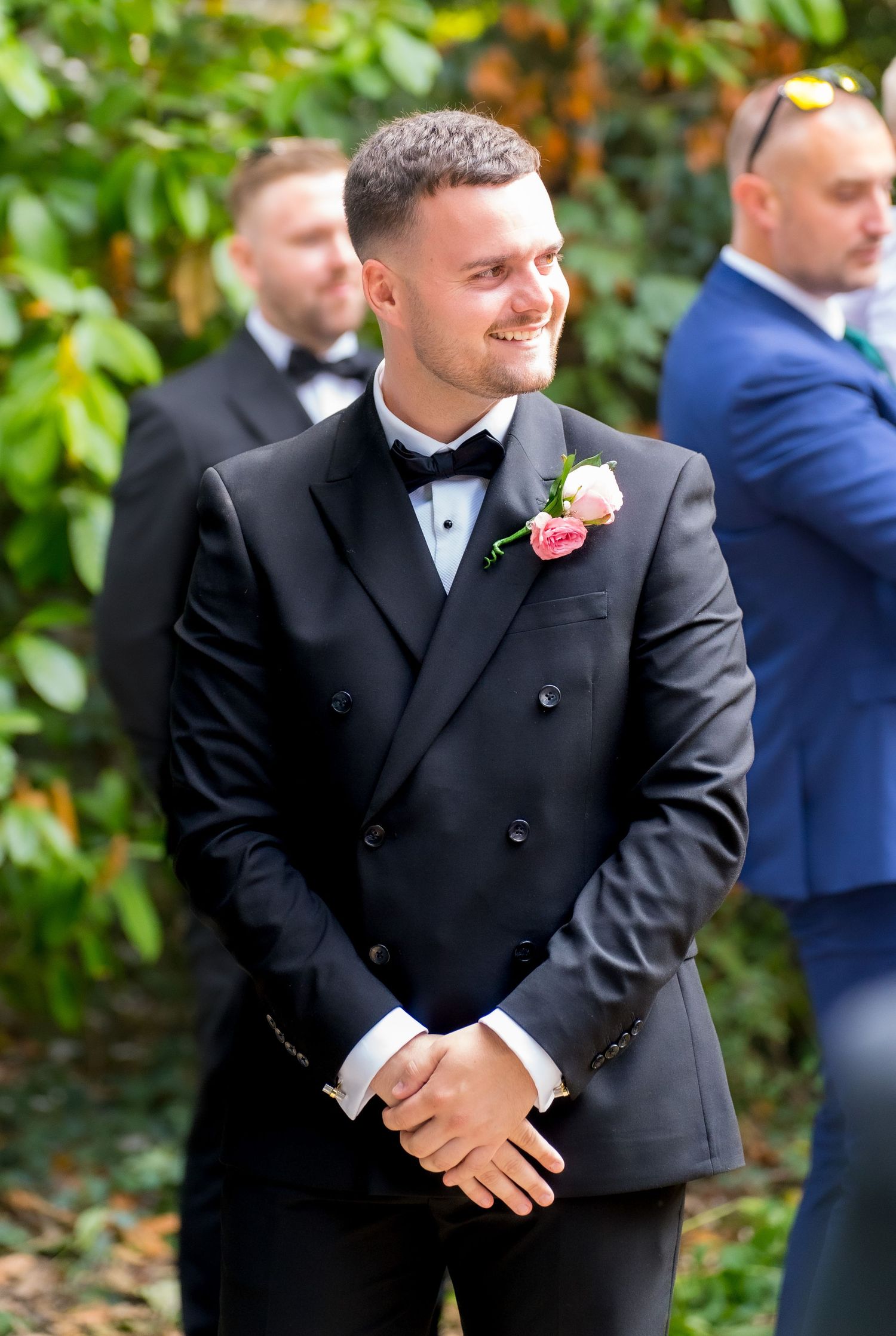 Handsome groom in black double-breasted tuxedo with pink boutonniere smiling at outdoor wedding ceremony.