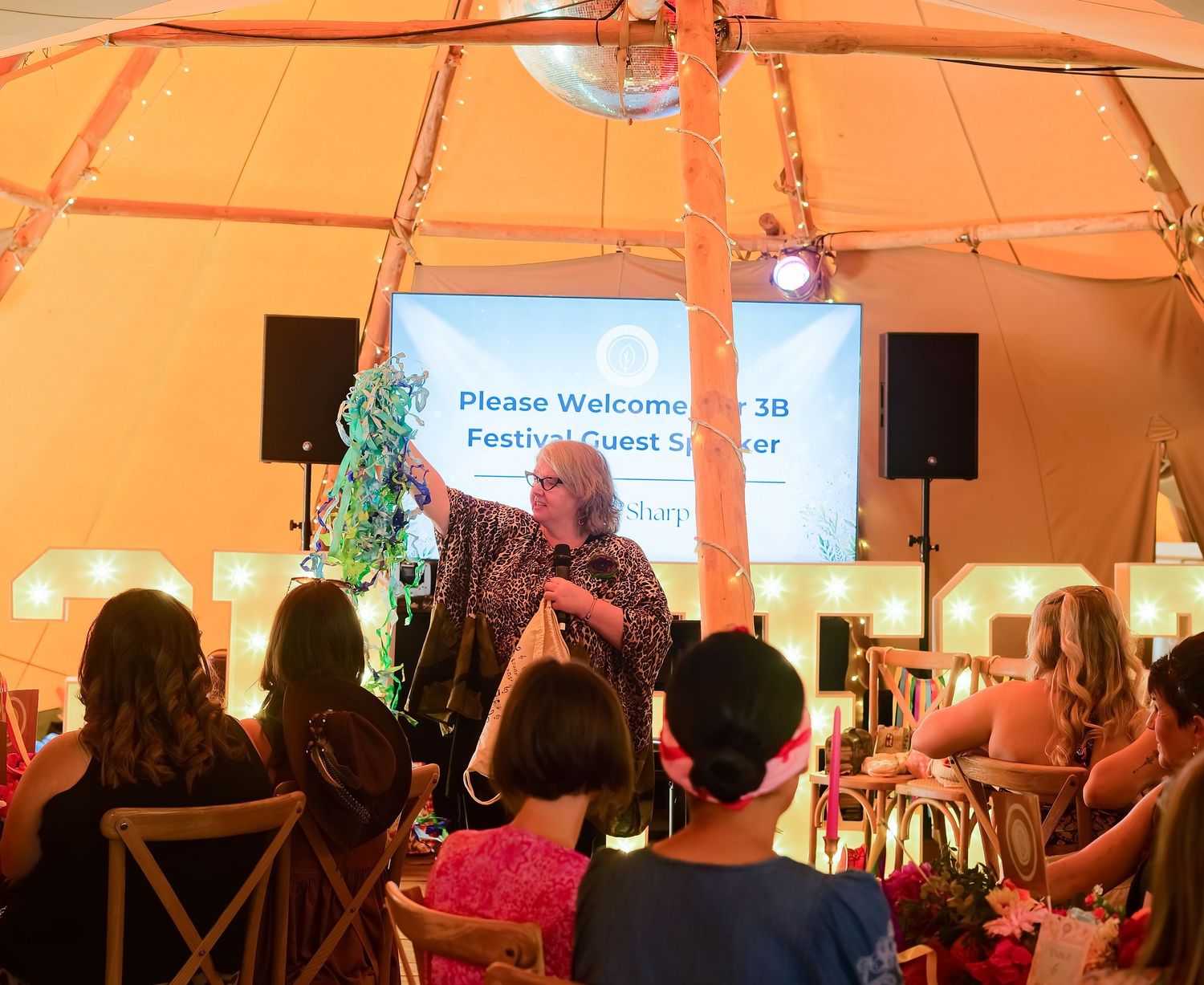 Speaker presenting at a 3B Festival Guest Speaker event inside a decorated tent with string lights and audience.