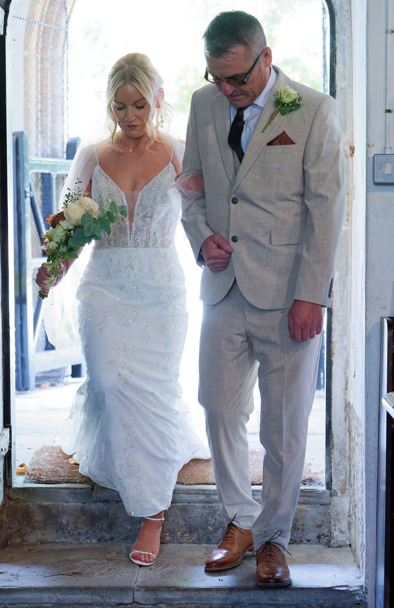Bride in white lace gown holding bouquet walks down steps with man in beige suit at wedding ceremony.