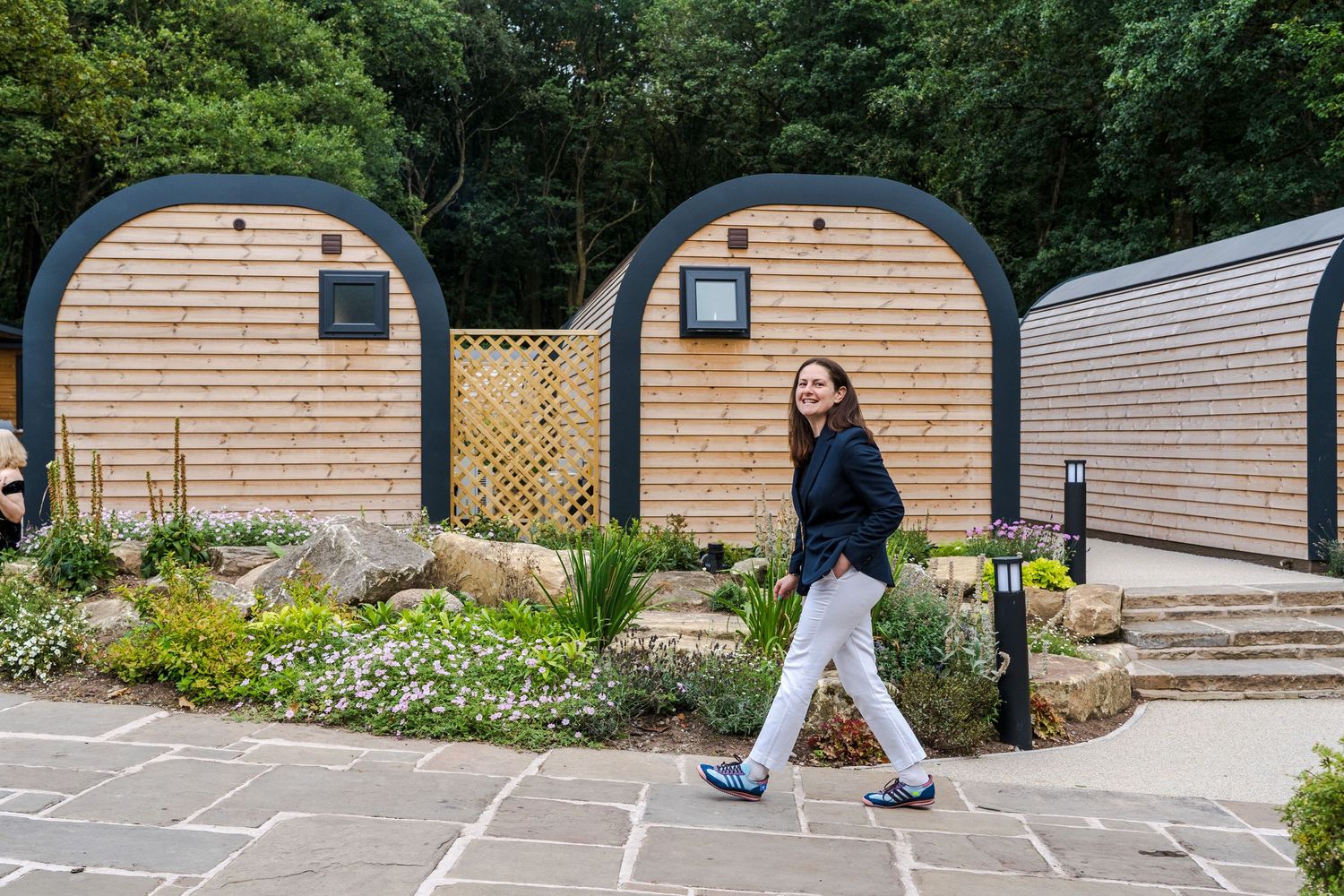 Woman walking past wooden glamping pods with arched roofs surrounded by landscaped gardens and lush greenery.