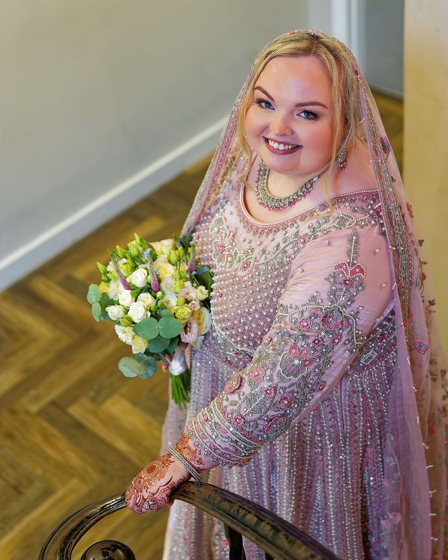 Bride in pink embroidered South Asian bridal gown with veil and henna, holding white bouquet on staircase.