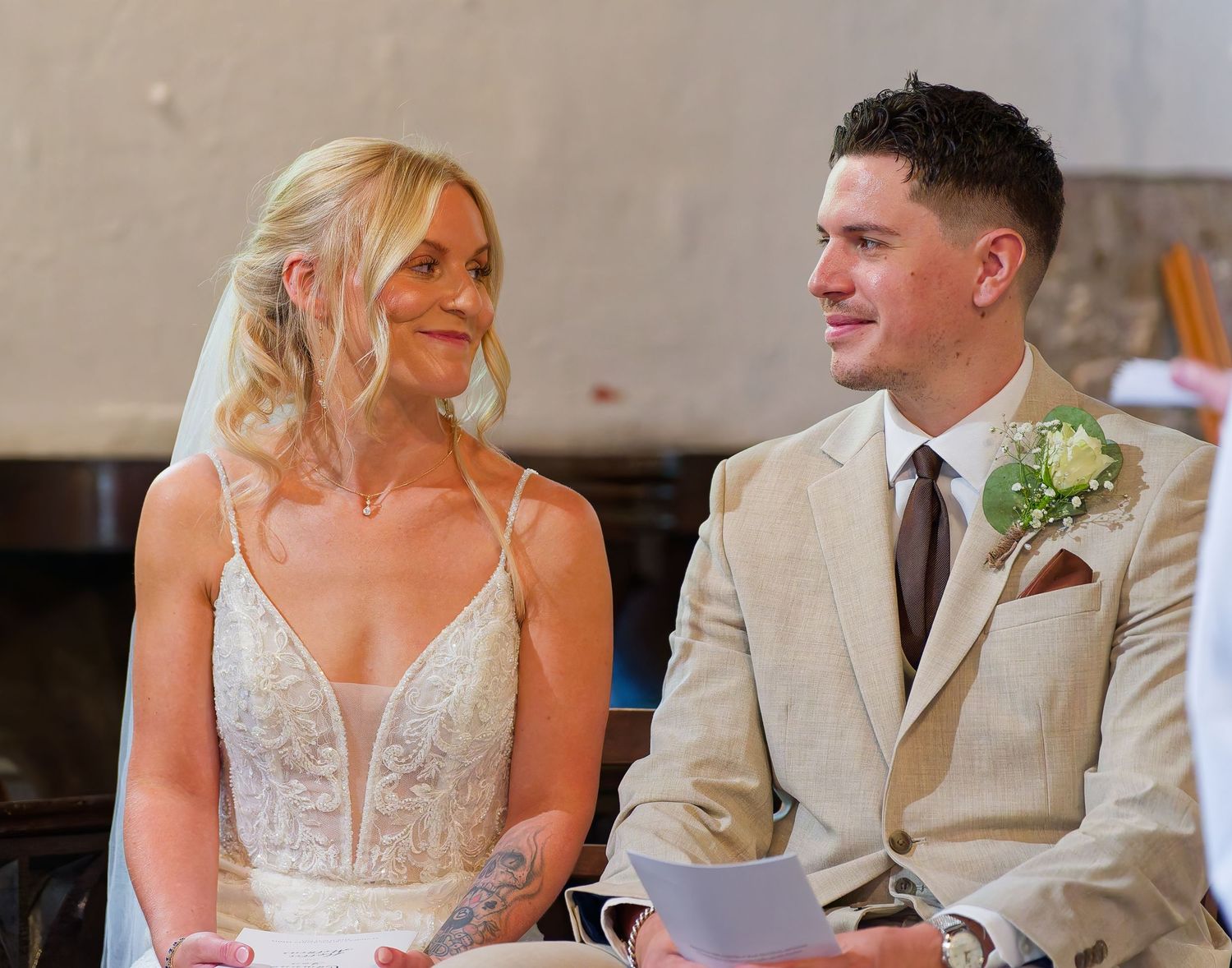 A smiling bride in a lace gown and groom in a tan suit with boutonniere share a joyful moment during their wedding ceremony.