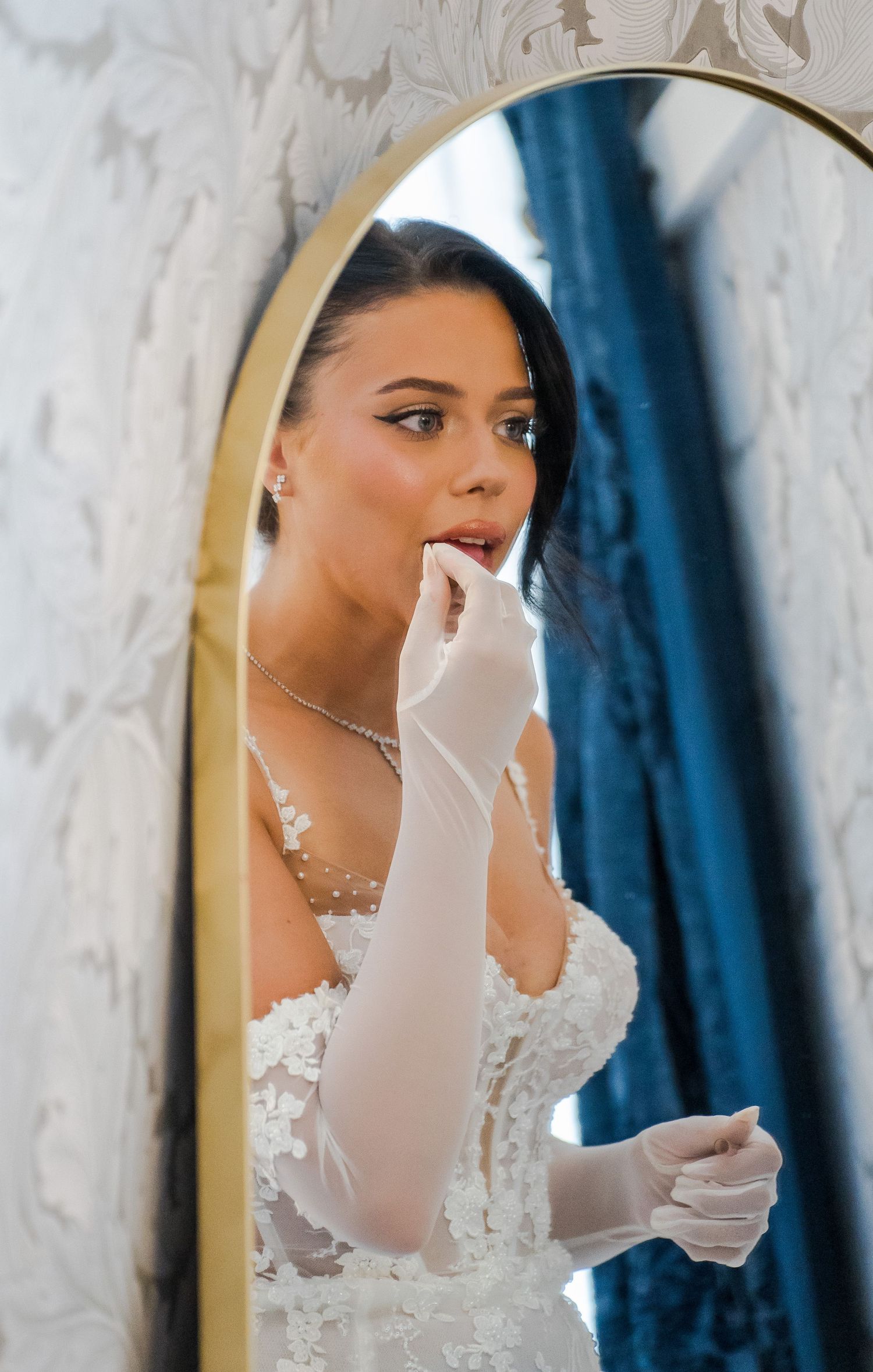 Bride in lace wedding dress and white gloves touches her lips while gazing into a gold-framed mirror.