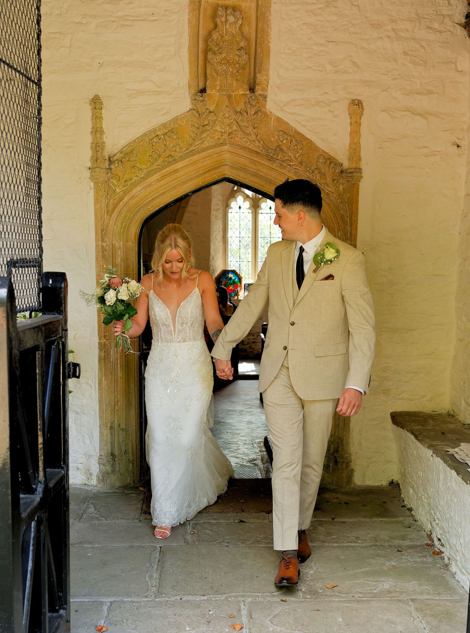 Bride and groom holding hands walking out of a stone church arch doorway, bride in white lace gown holding bouquet.