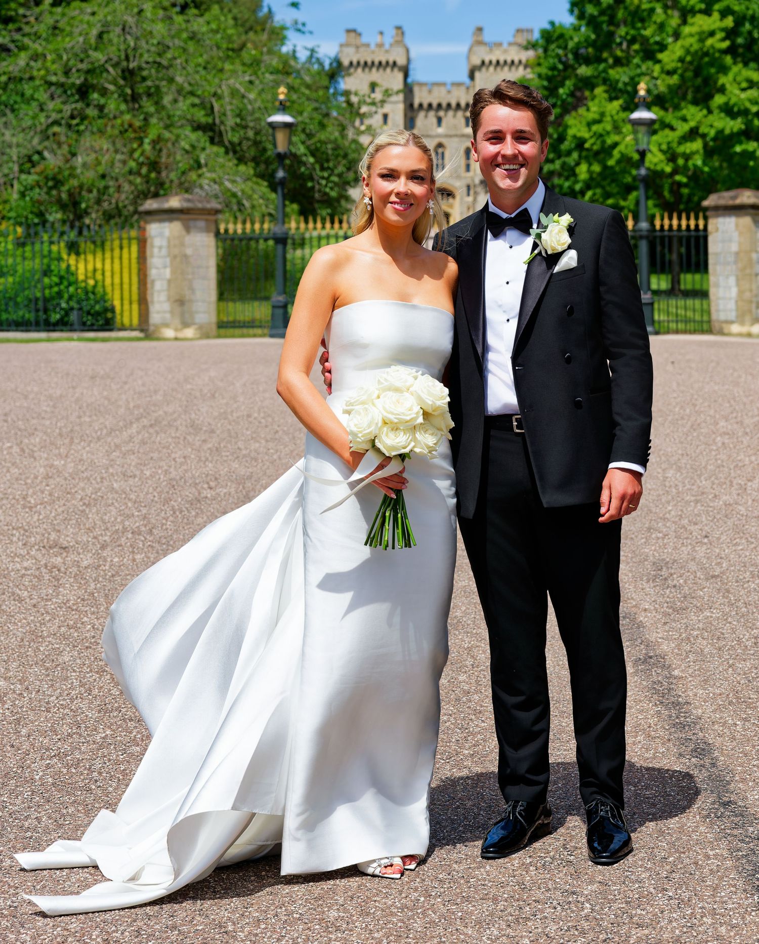 Bride in strapless white gown holding white roses bouquet poses with groom in black tuxedo outside Windsor Castle.