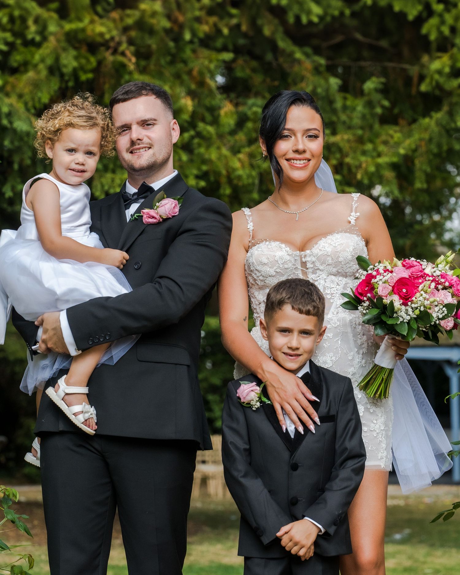Bride and groom in formal wedding attire pose outdoors with two young children, holding a pink and white bouquet.