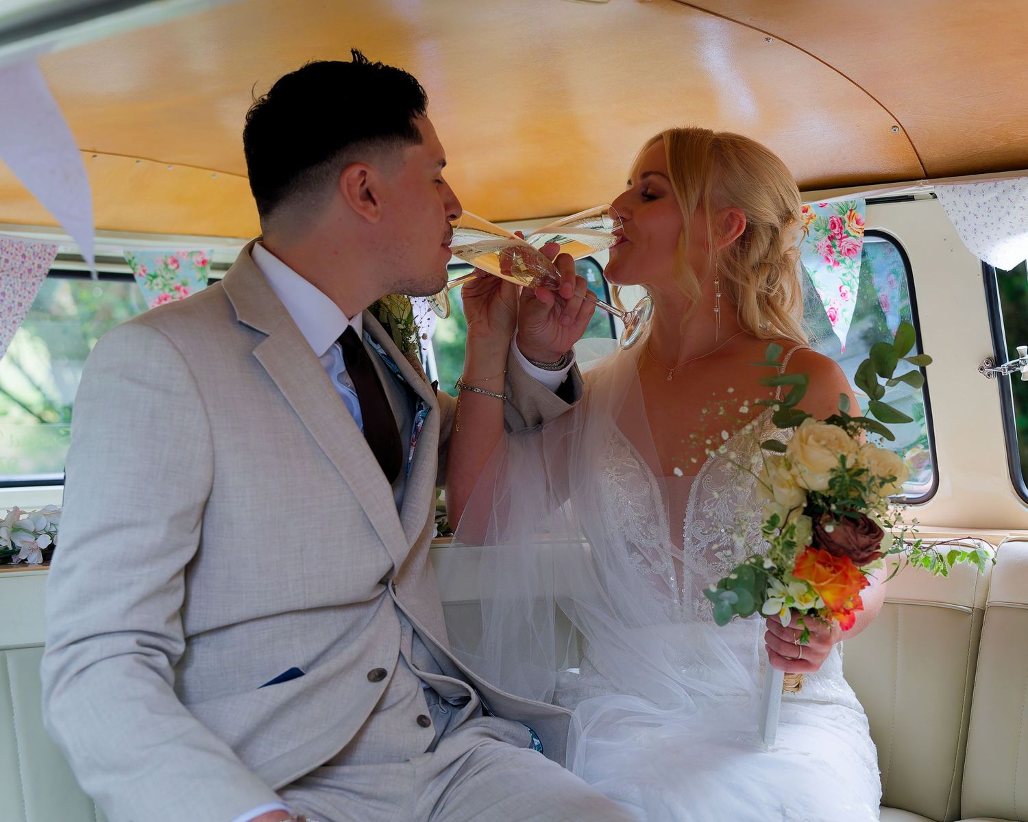 Newlywed couple sharing a romantic moment in a vintage VW campervan, bride holding bouquet, groom in grey suit.