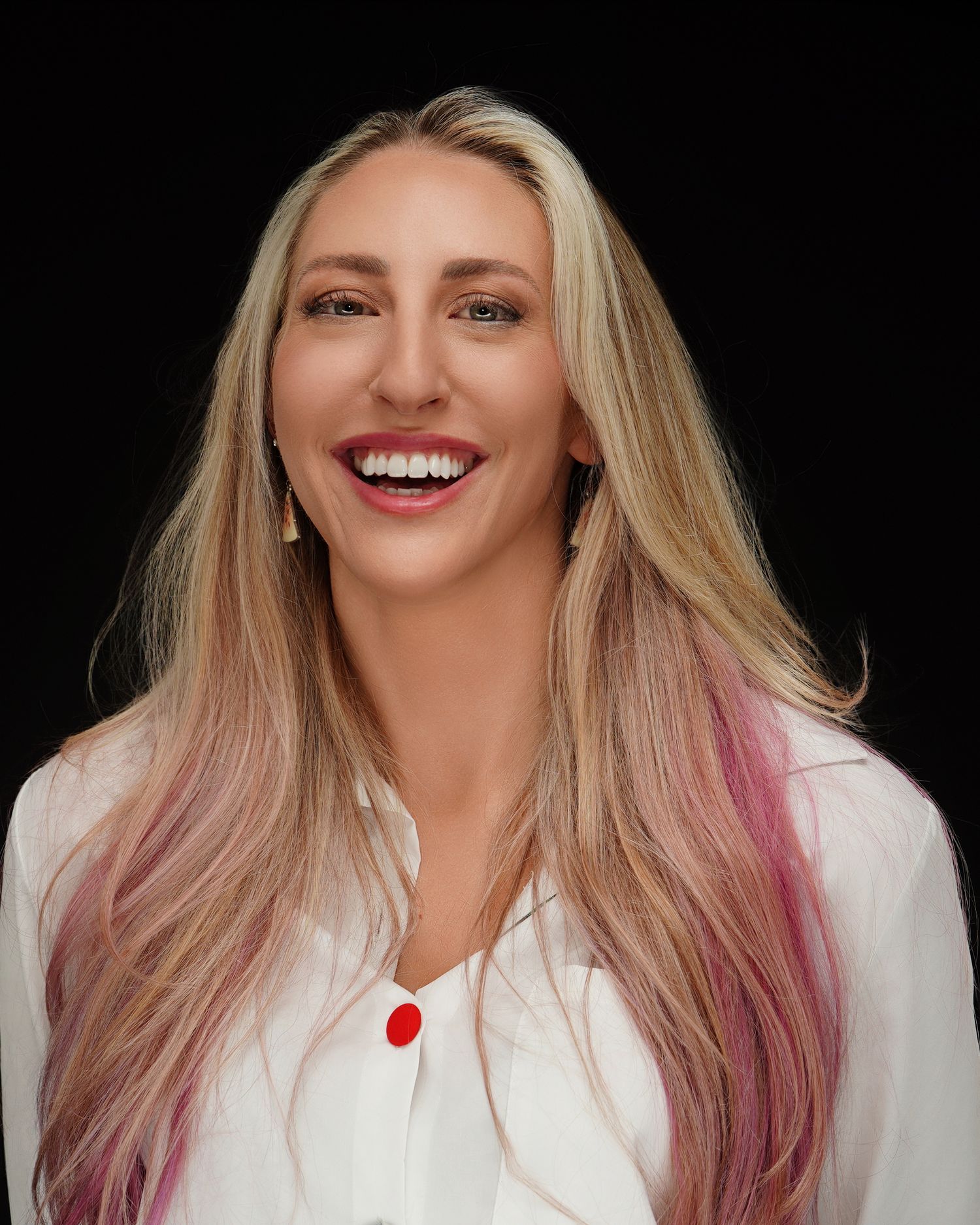 Professional portrait of a person with flowing blonde and pink hair wearing a white top against a dark background.
