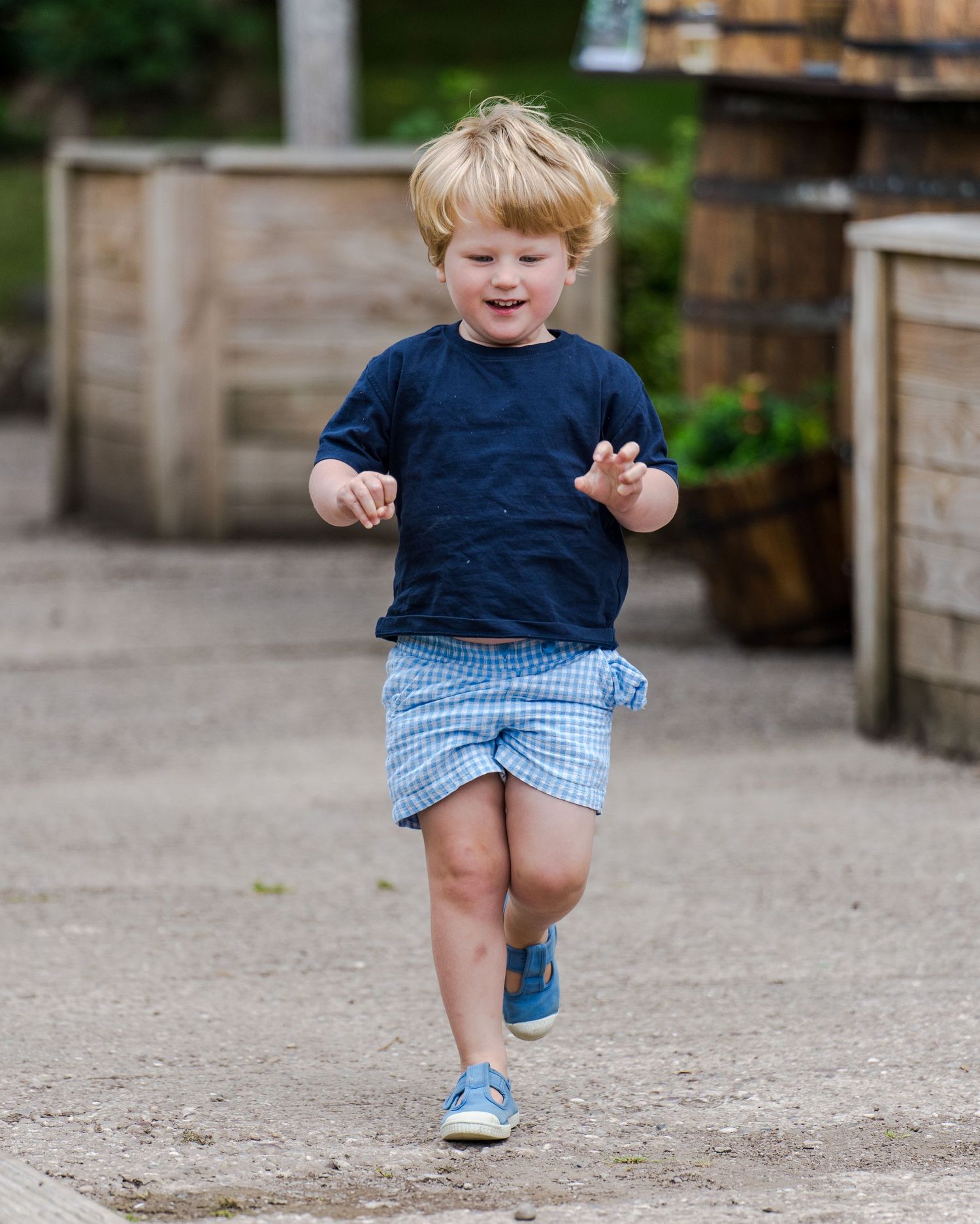 A joyful child runs on a path in a navy blue shirt and checkered blue shorts with light blue sneakers.