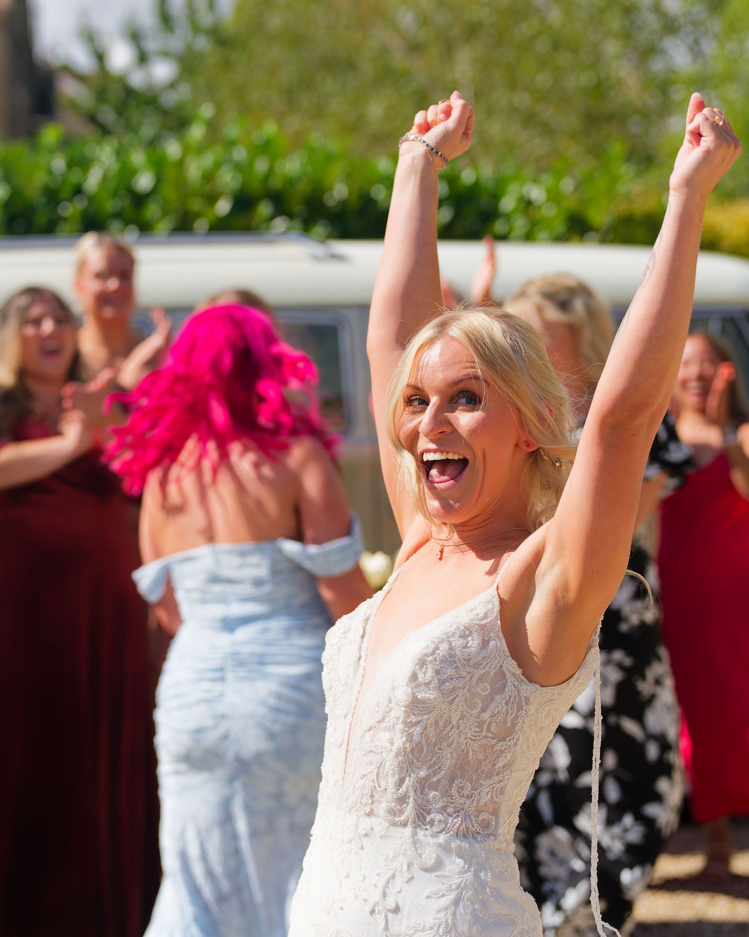 Excited blonde bride in lace gown celebrating with arms raised at outdoor wedding.