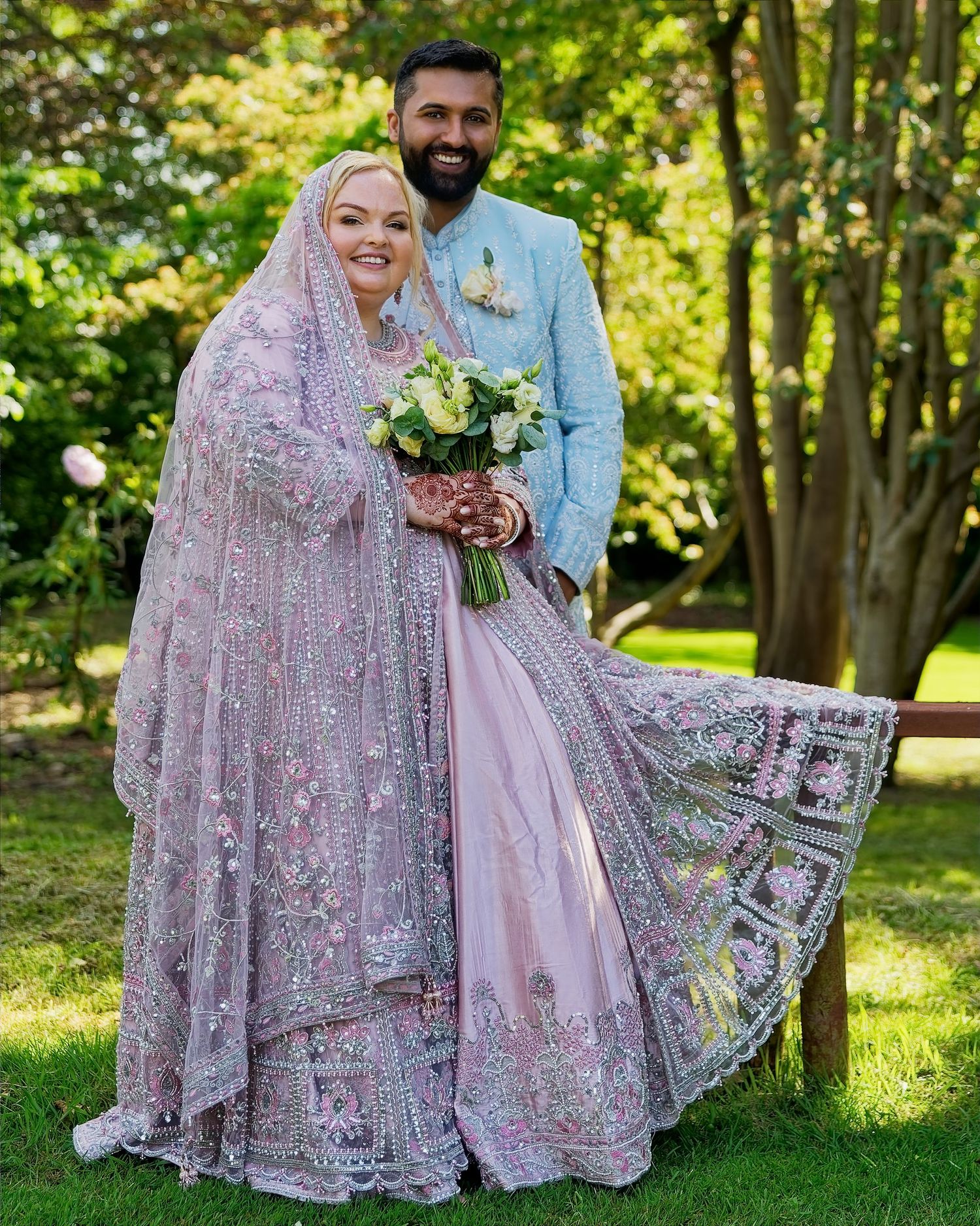 Couple in traditional South Asian wedding attire, bride in lavender lehenga and groom in blue sherwani, posing outdoors.