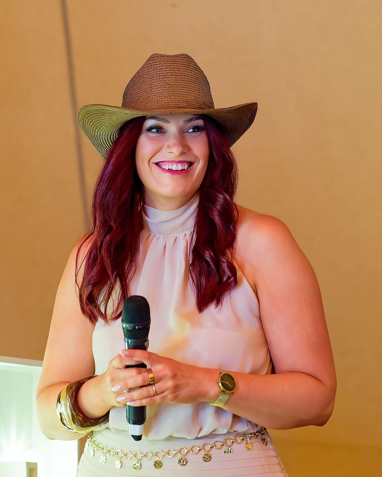 Woman with red hair and cowboy hat smiling while holding a microphone at an event.