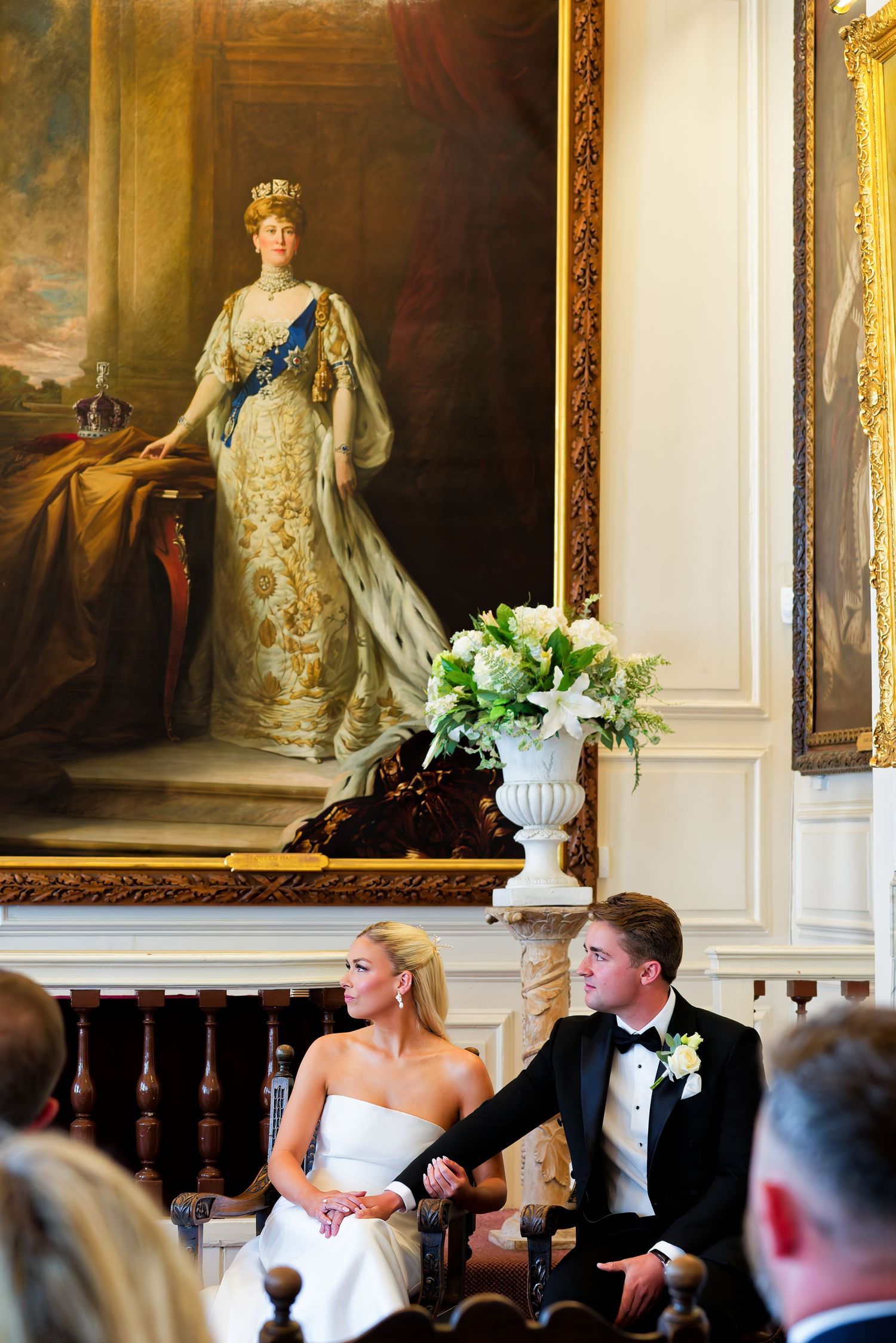 Bride and groom seated at elegant wedding ceremony beneath a grand royal portrait in an ornate historic venue.