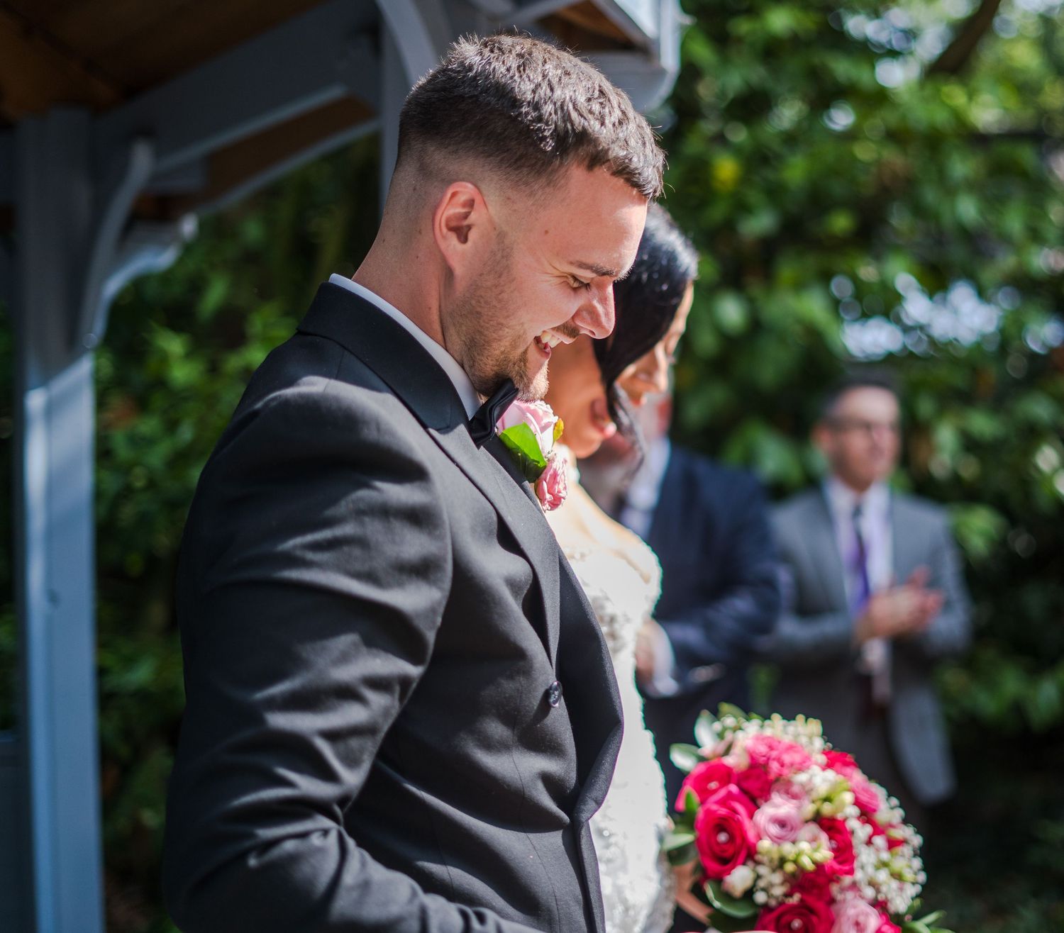 Emotional groom in black tuxedo bows head during outdoor wedding ceremony with pink floral boutonniere.