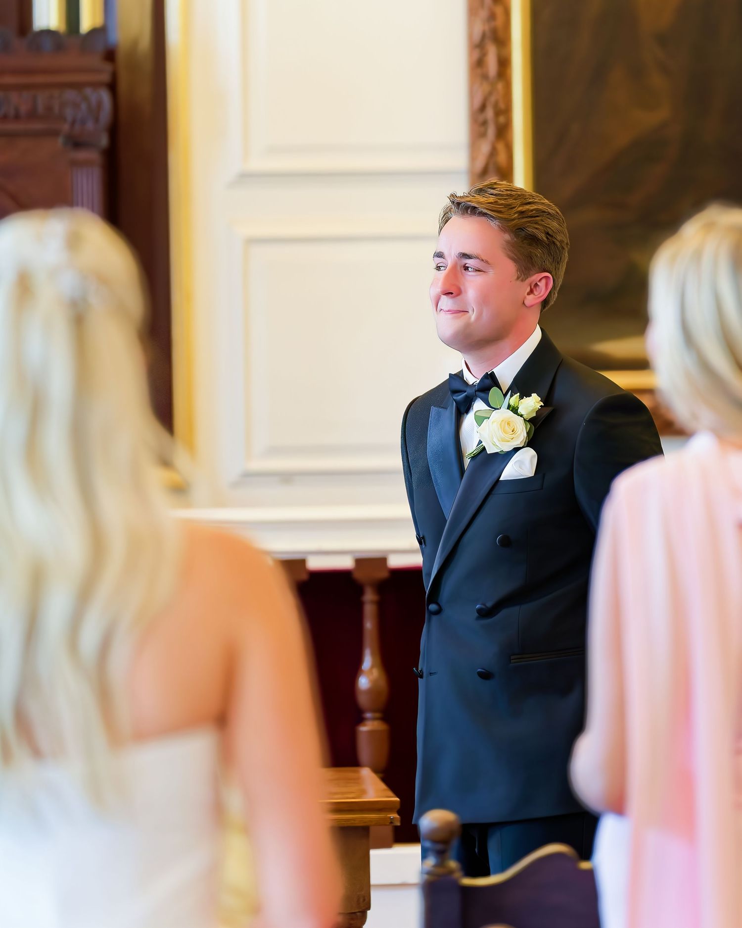 Emotional groom in black tuxedo watches bride approach during elegant wedding ceremony indoors.
