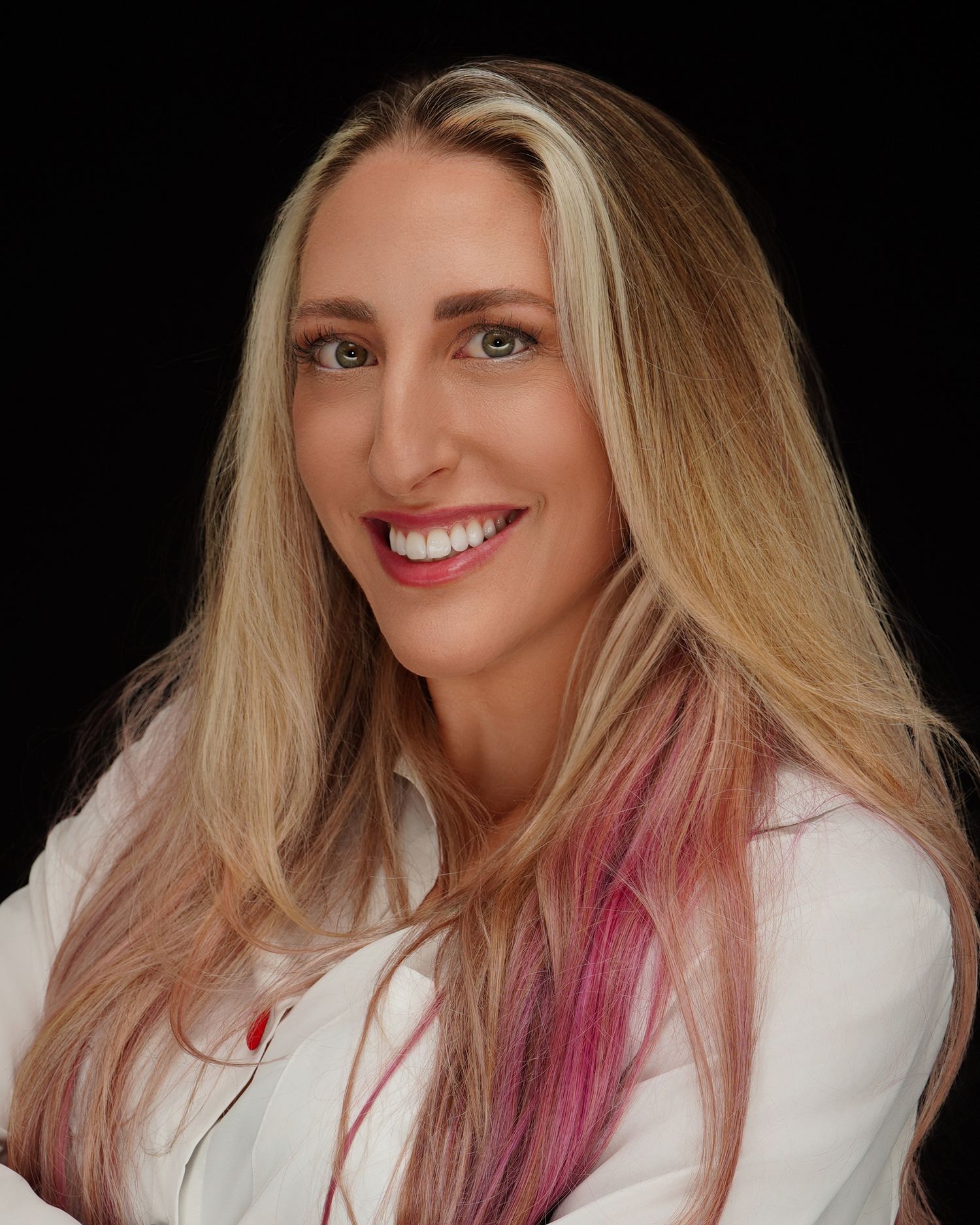 Professional headshot of a smiling blonde woman with pink highlights wearing a white blazer against a black background.