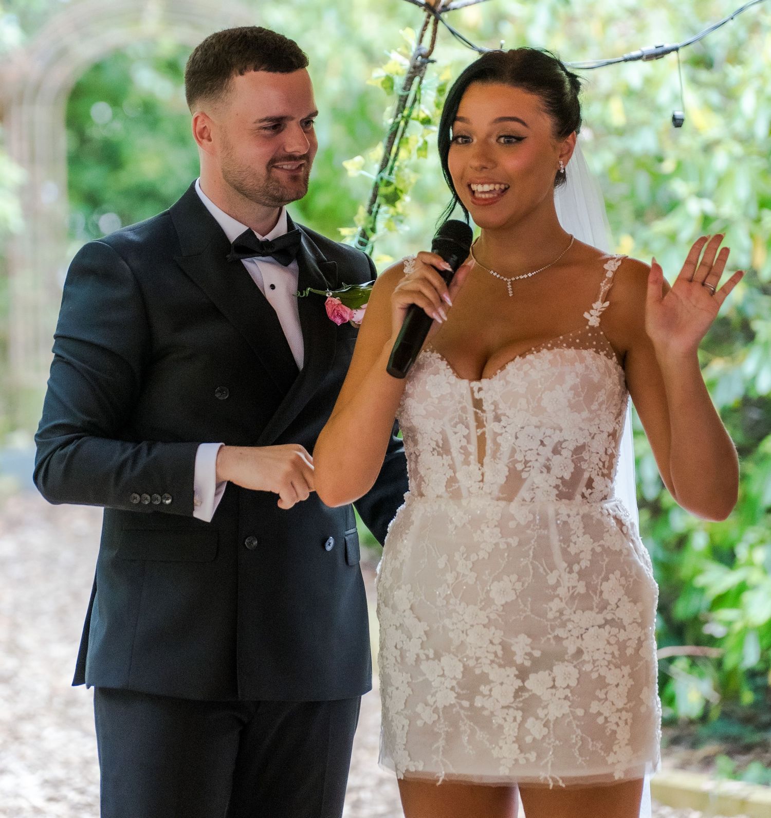 Bride in short lace wedding dress and groom in tuxedo speaking at their wedding ceremony with microphone.