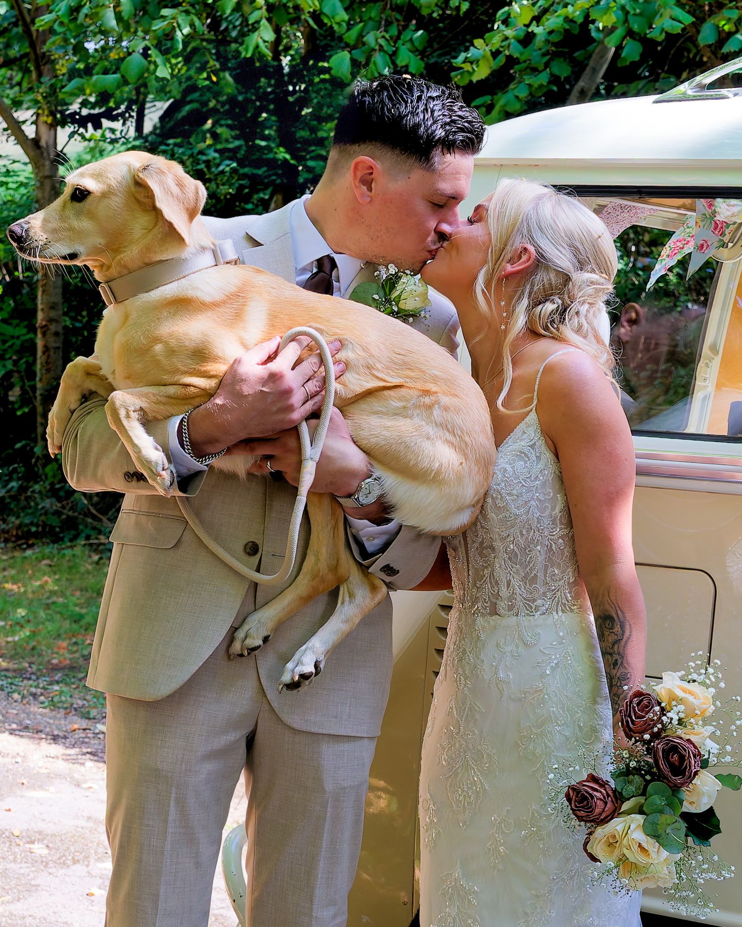 Bride and groom kissing while holding a yellow Labrador dog on their wedding day near a vintage van.