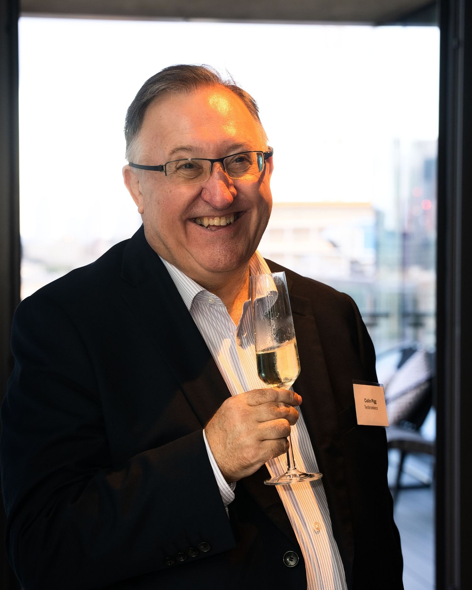 Smiling man in glasses and a dark blazer holds a champagne flute at an evening event with a city view behind him.