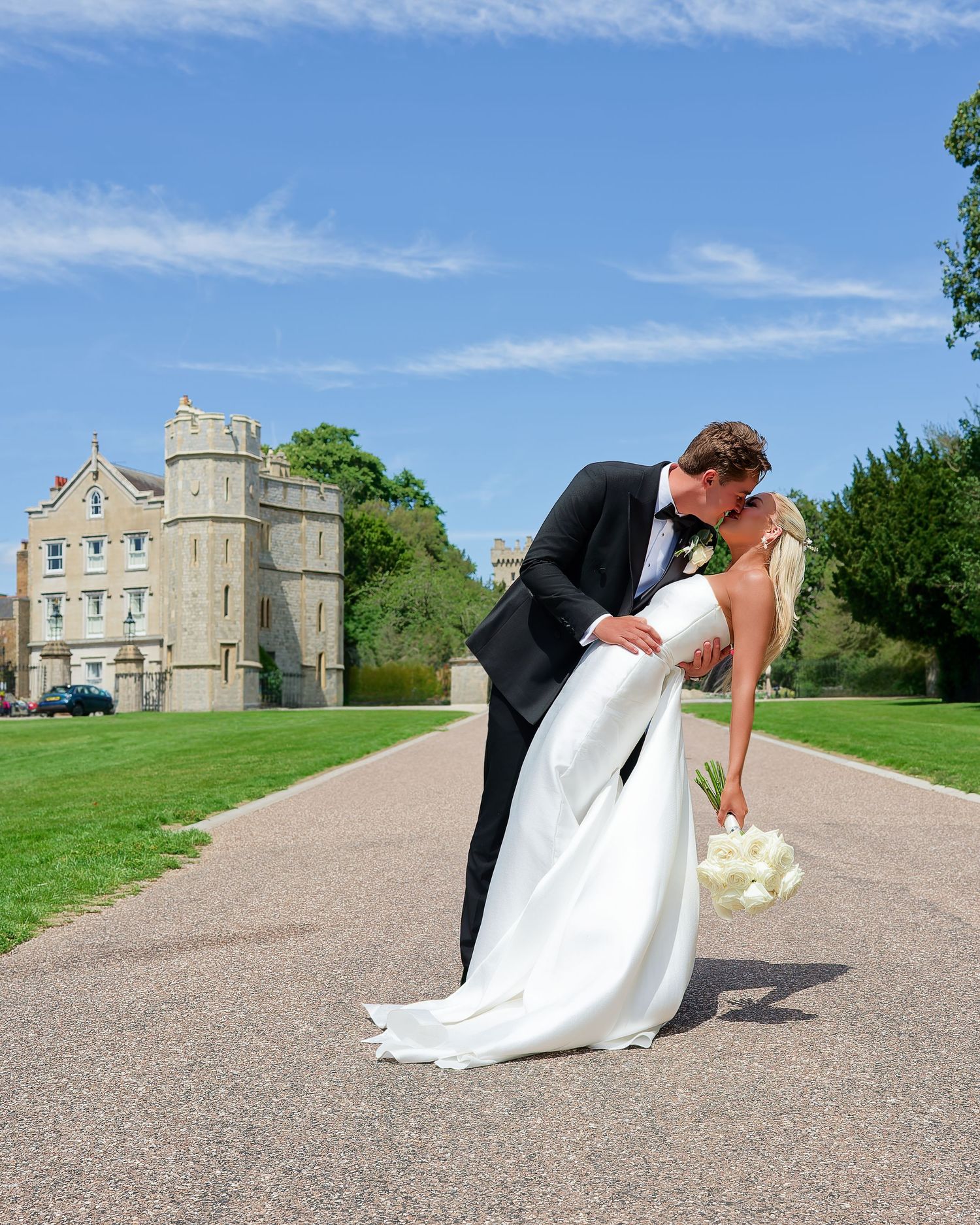Groom dipping bride for a kiss on a scenic castle driveway under a bright blue sky.