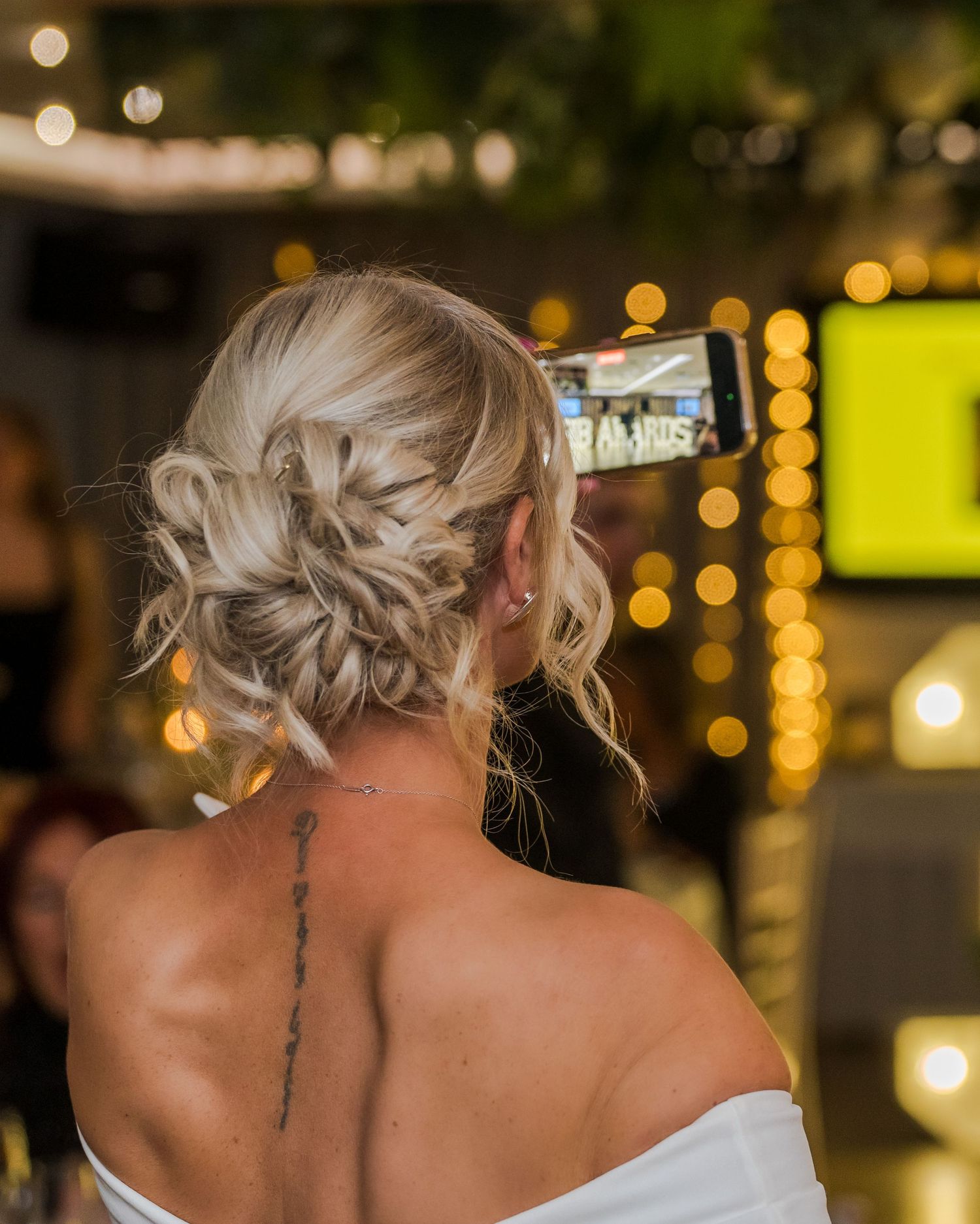 Bride with elegant braided updo hairstyle and neck tattoo in off-shoulder dress at wedding reception.