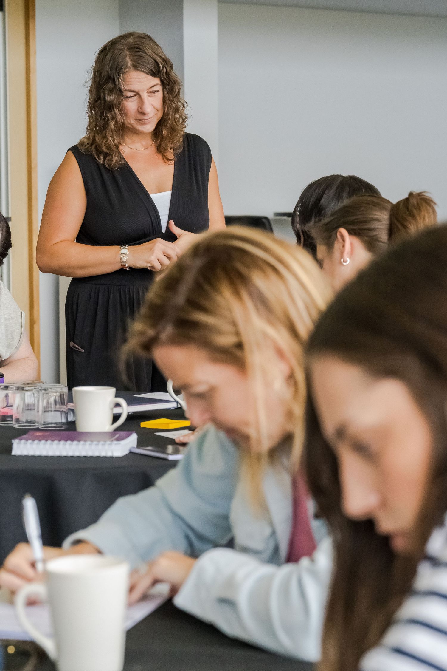 A woman in a black dress stands and observes while seated participants take notes at a table with coffee mugs during a workshop.