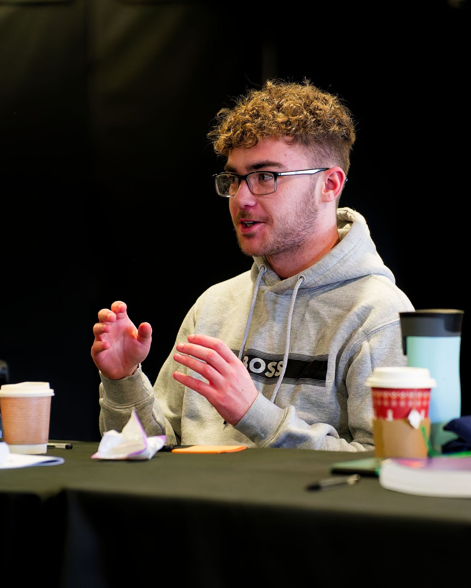 Young man with curly hair and glasses gestures while speaking at a table with coffee cups, a water bottle and notes.