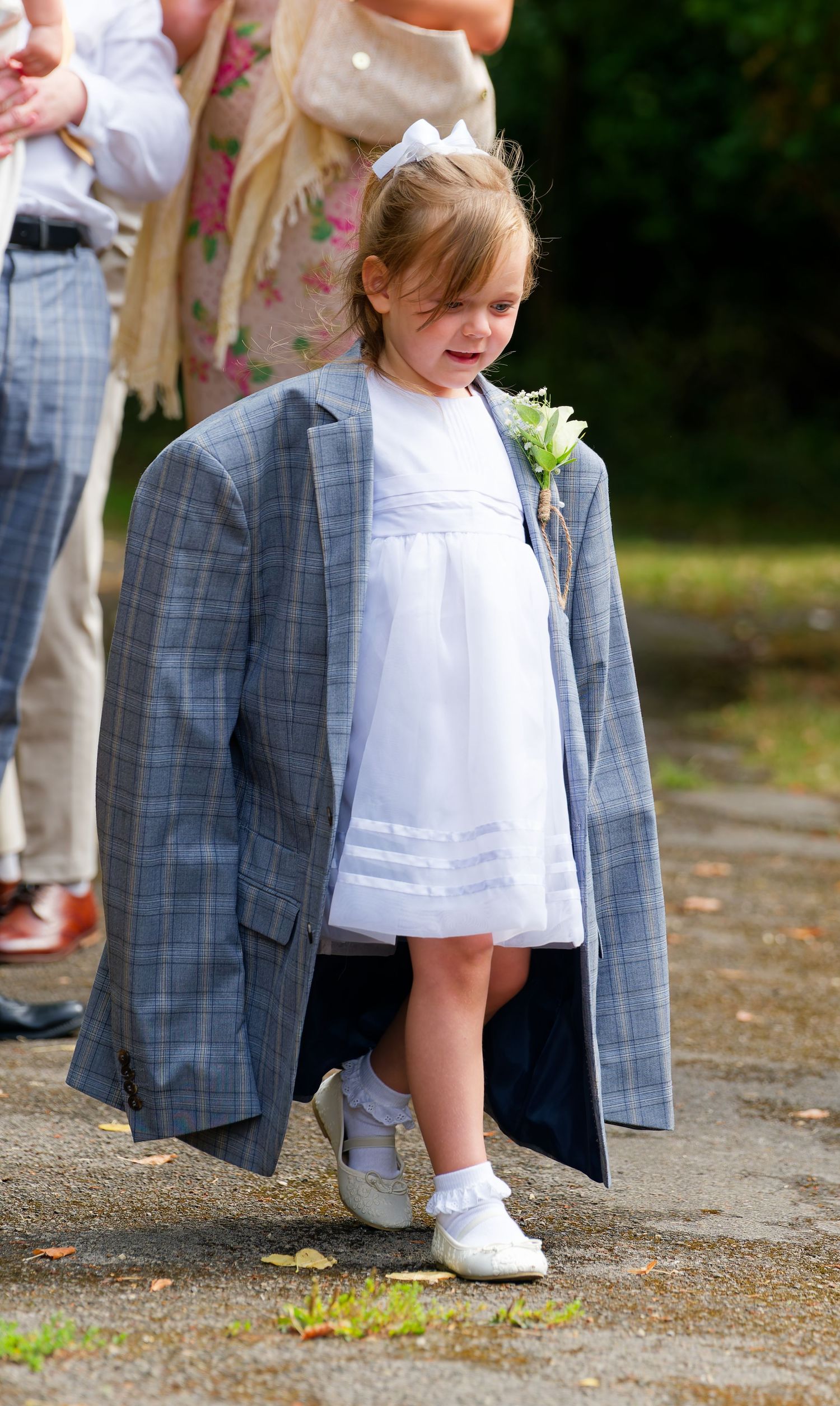 Young flower girl in white dress wearing oversized gray blazer at a wedding ceremony outdoors.