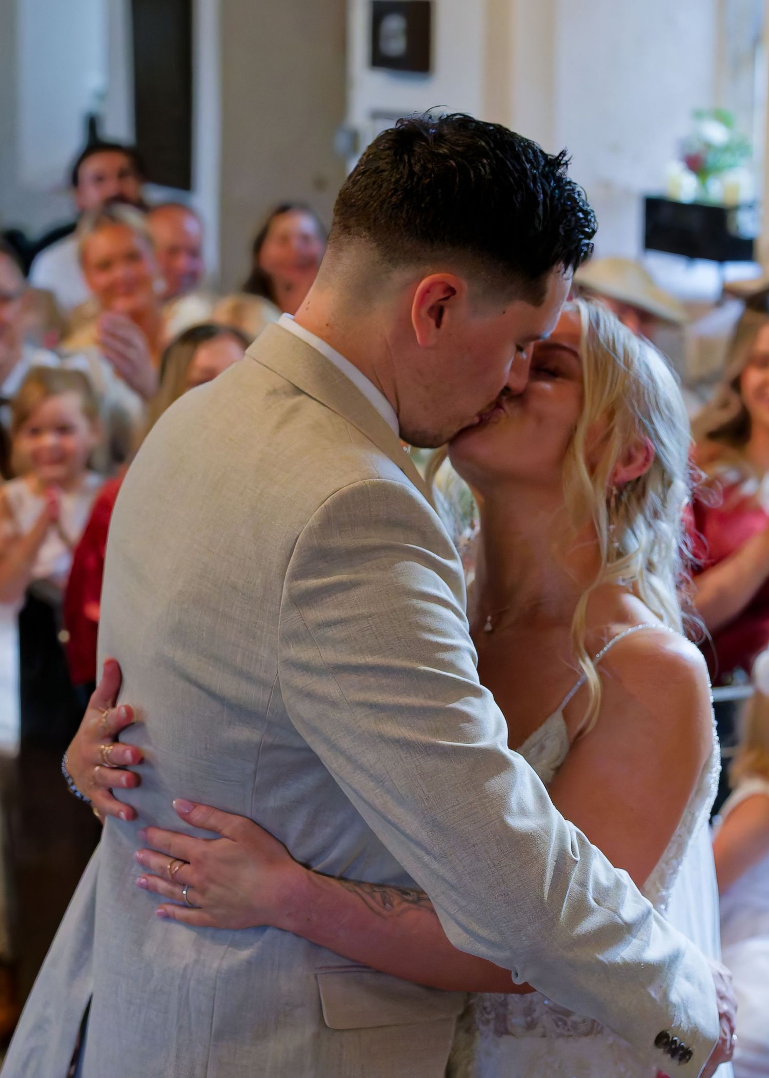 Newlywed couple sharing their first kiss at wedding ceremony with guests applauding in background.