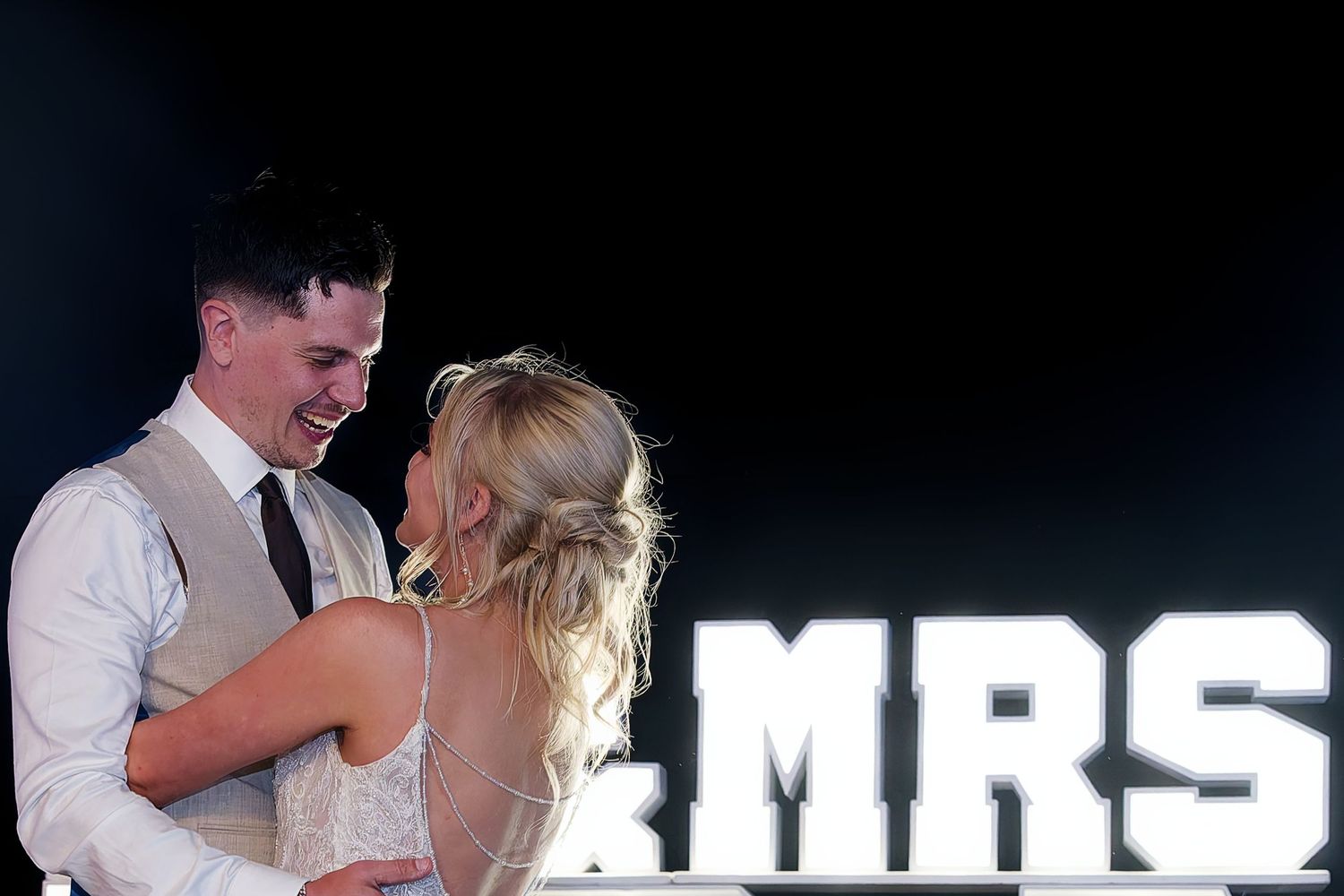 Bride and groom share first dance in front of illuminated MRS marquee letters at elegant wedding reception.